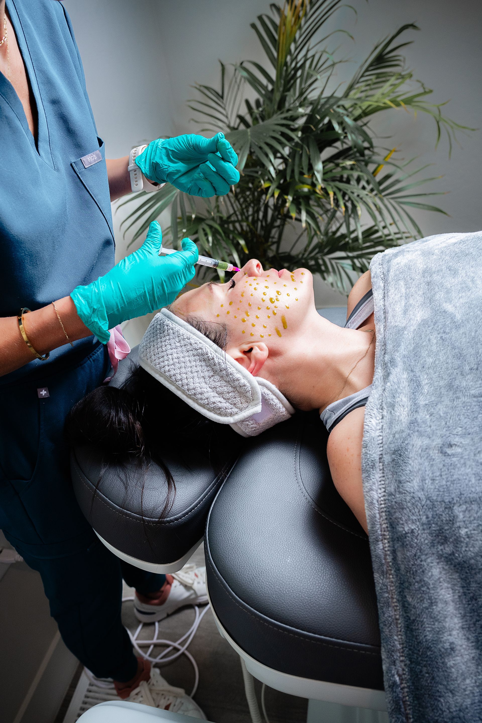 Gloved clinician applies facial treatment to a client reclining in a spa chair.