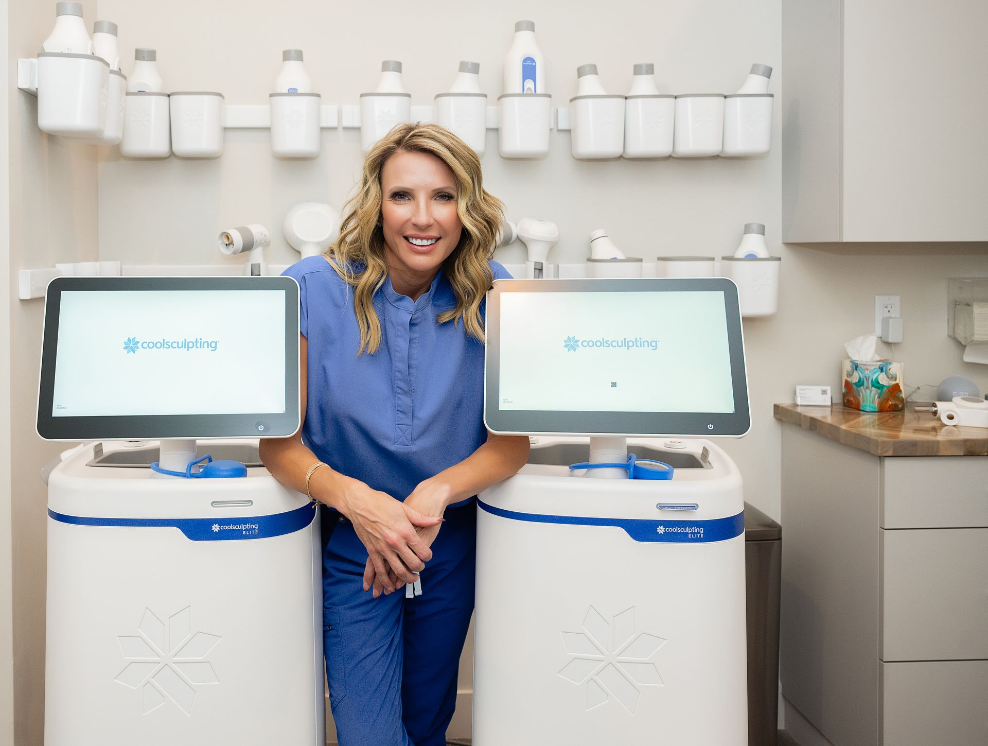 A woman in a blue scrub is standing next to two computer monitors.