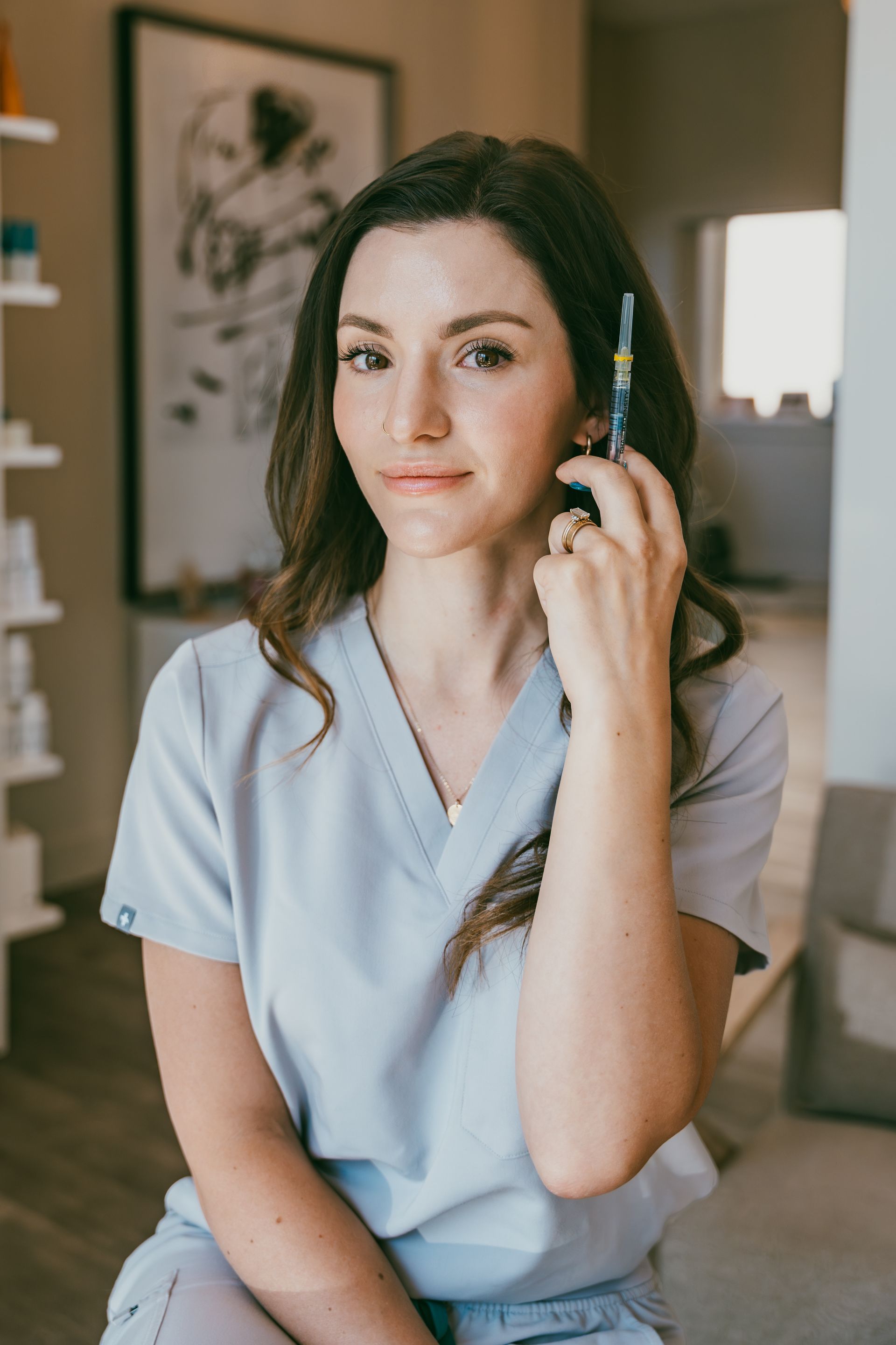 Woman in blue scrubs holding a syringe, looking at the camera. She is indoors.