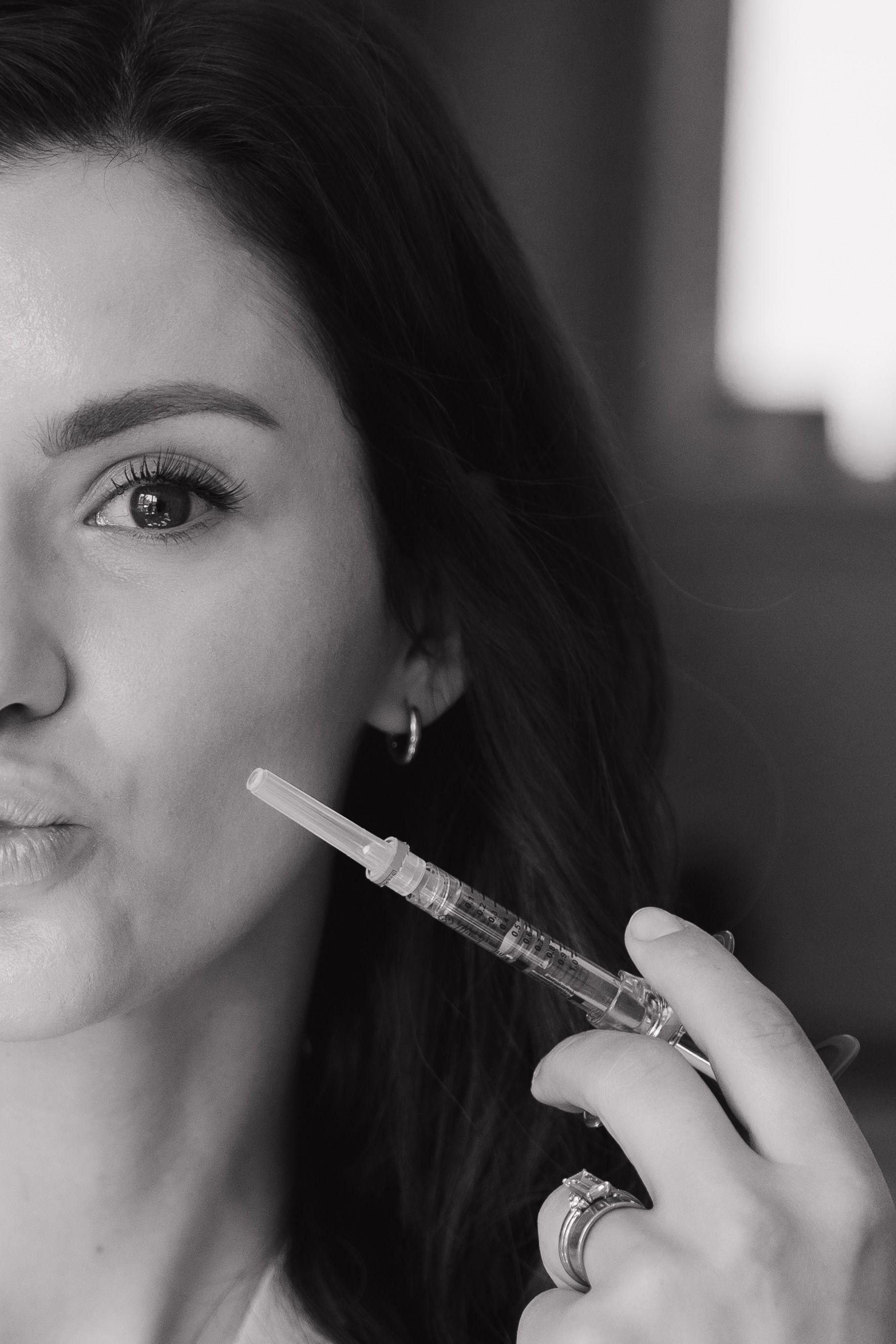 Black and white photo of a woman holding a syringe near her face, looking towards the camera. She has dark hair and is wearing rings.