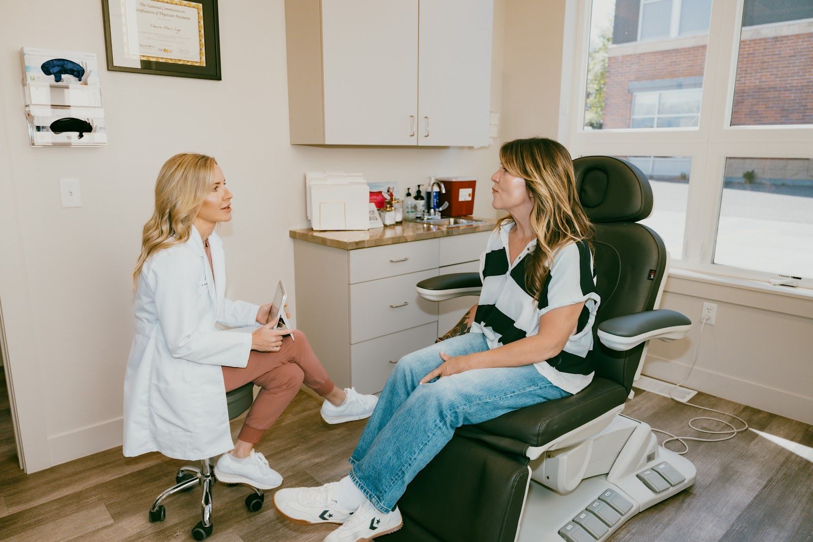 Doctor and patient in a medical office. Doctor in white coat sits, talking to patient seated in a chair, sunlight through windows.