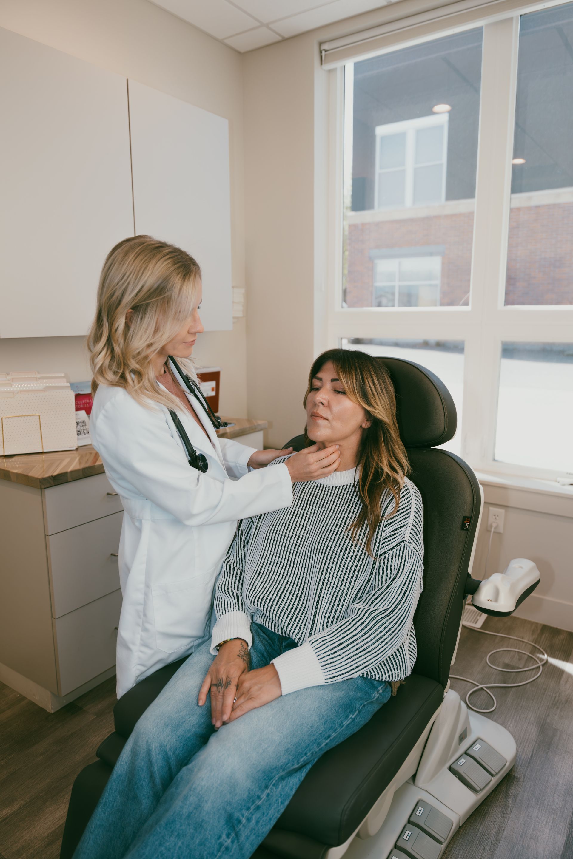 A female doctor examines a patient's neck in a medical office. The patient sits in a chair, and the doctor wears a white coat.