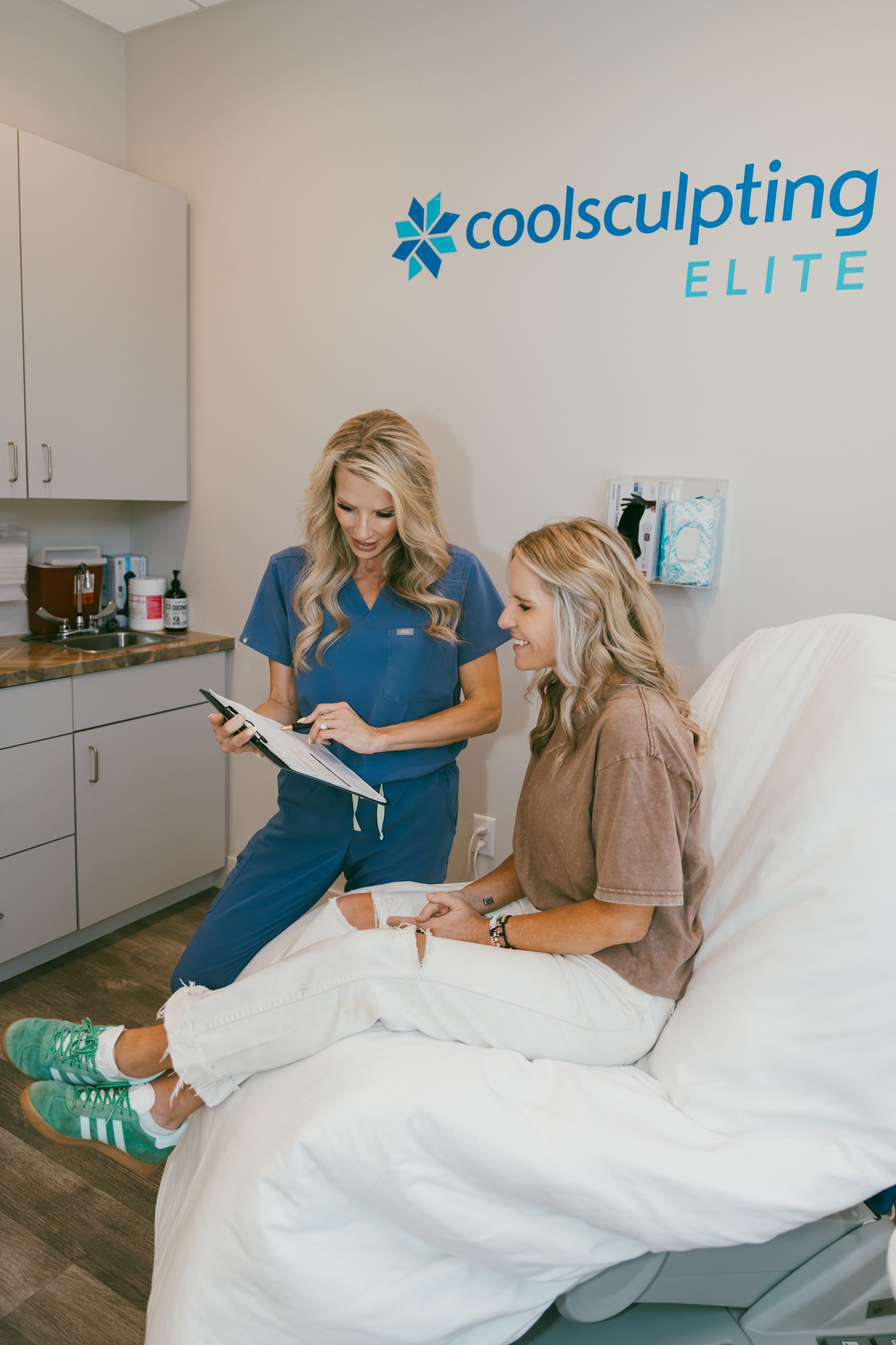 A medical professional in blue scrubs reviews paperwork with a patient seated on an examination table. The office features a