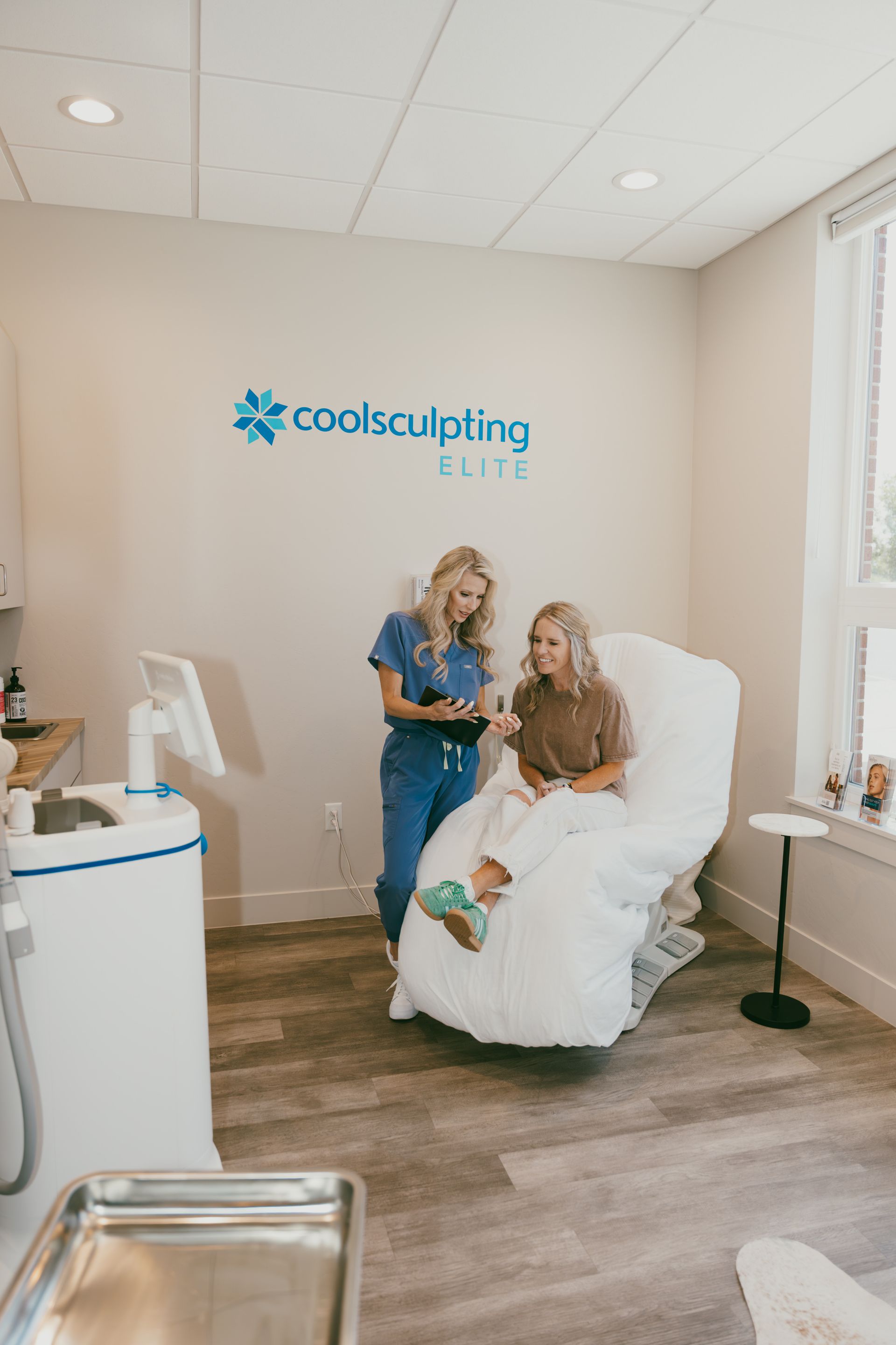 A medical professional in blue scrubs consults with a client seated on a treatment chair in a clinic. The room features a