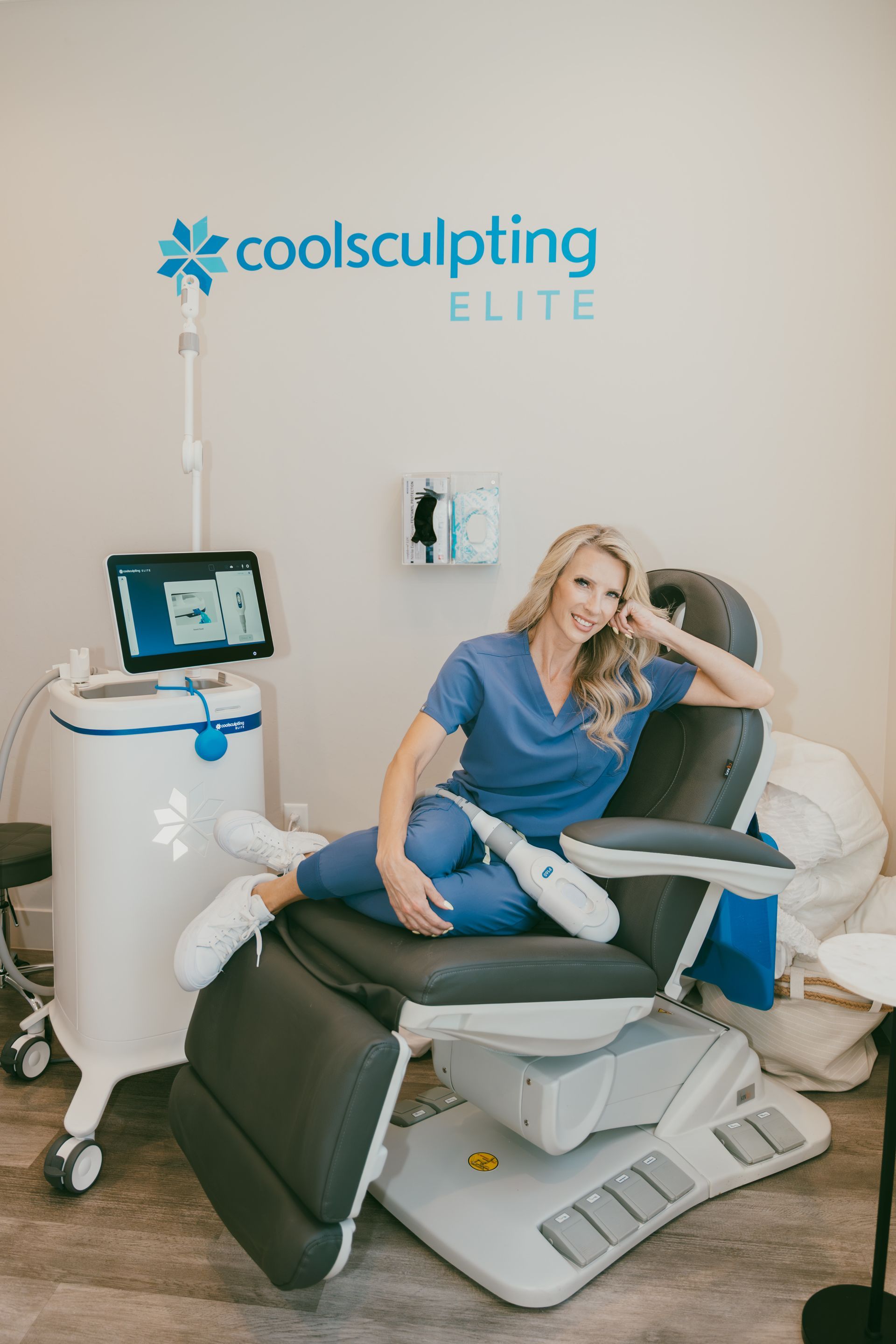 A blonde woman in blue scrubs poses in a treatment chair with CoolSculpting equipment. She smiles in a medical office setting.