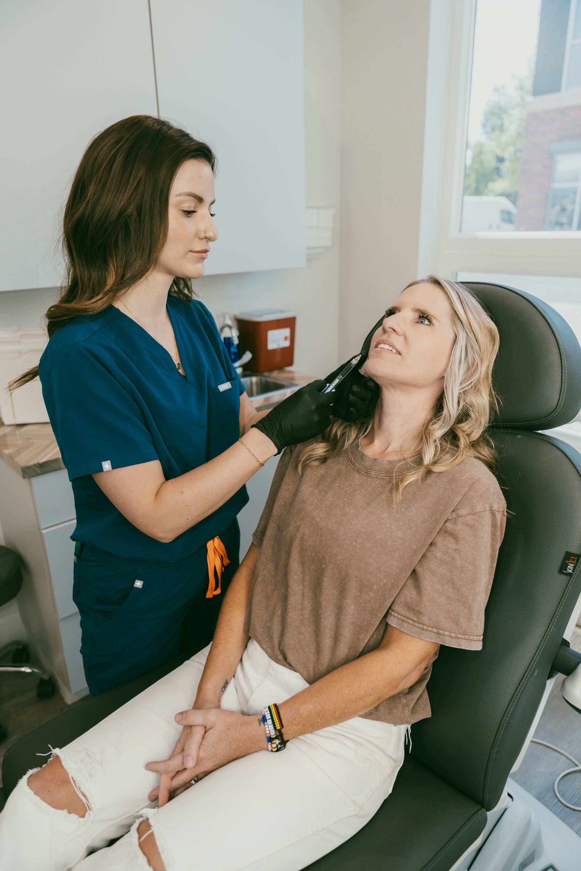 A medical professional in blue scrubs examines a seated woman's face. The woman is in a chair, anticipating treatment.