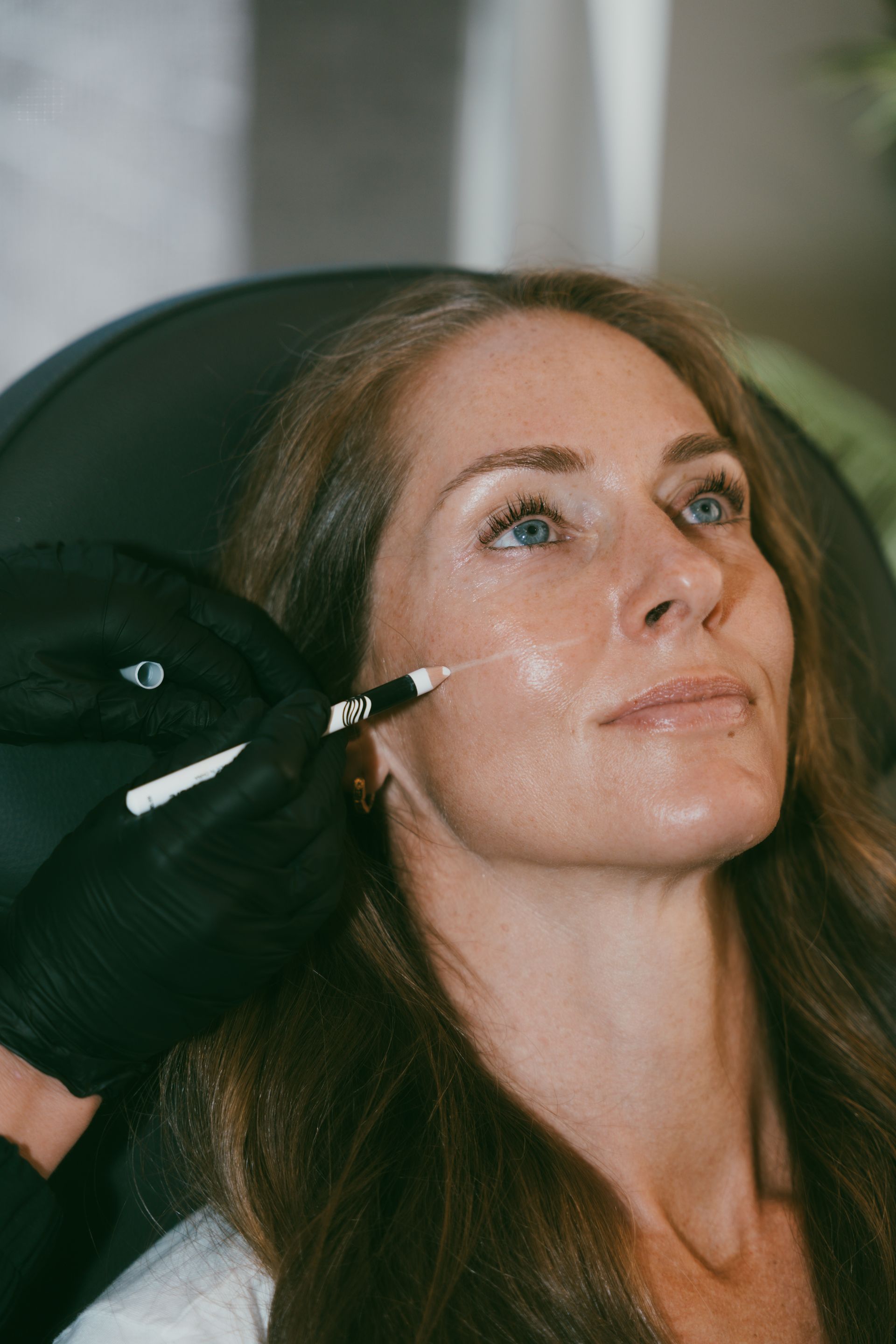 Woman receiving a cosmetic injection in her cheek. A gloved hand holds a syringe. Indoor setting, soft lighting.