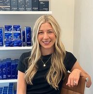 Woman with long blonde hair, smiling in front of a shelf of blue skincare products.