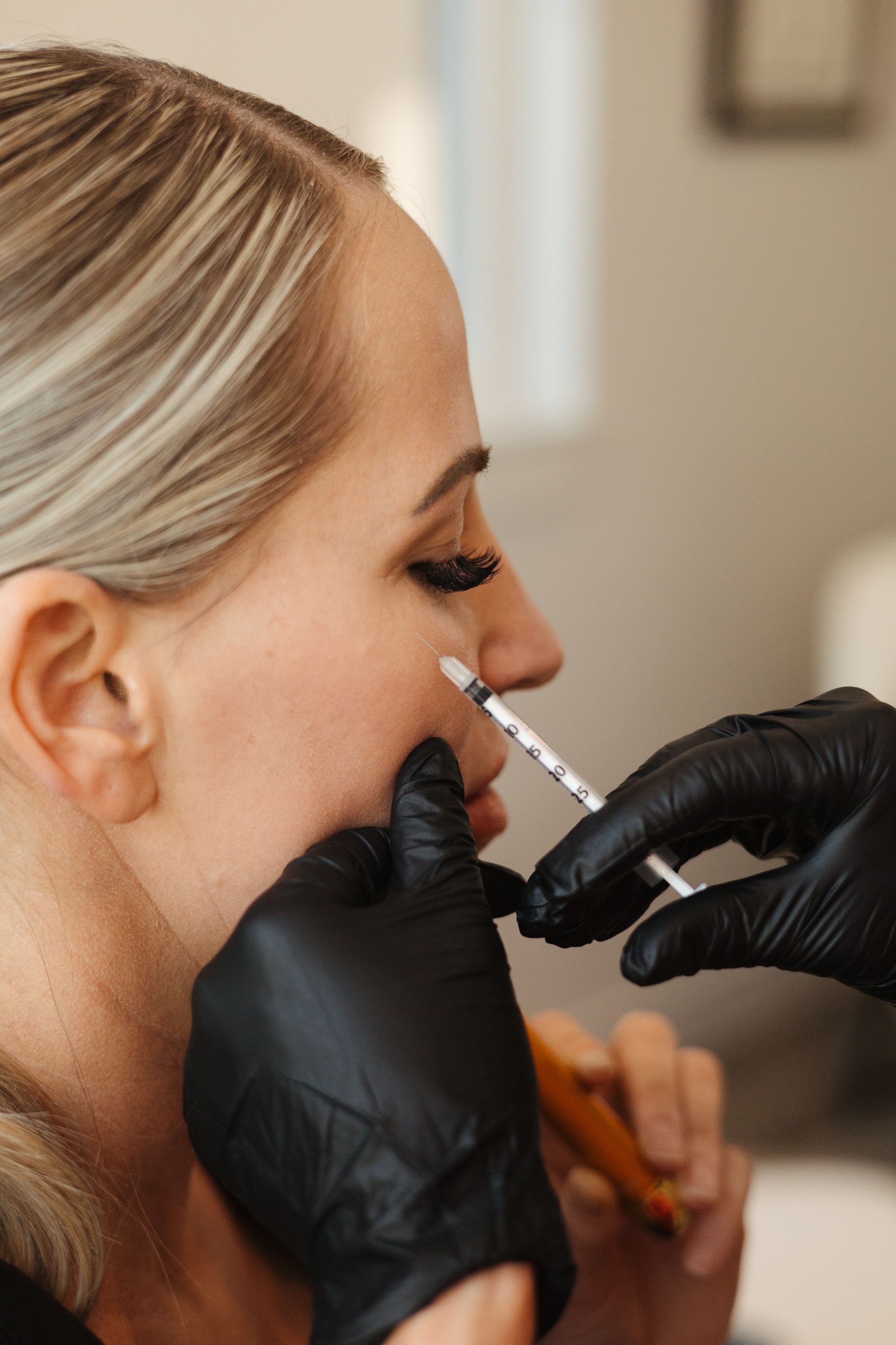 A woman receives a facial injection from a person wearing black gloves. Blonde hair frames her face in a beauty clinic setting.