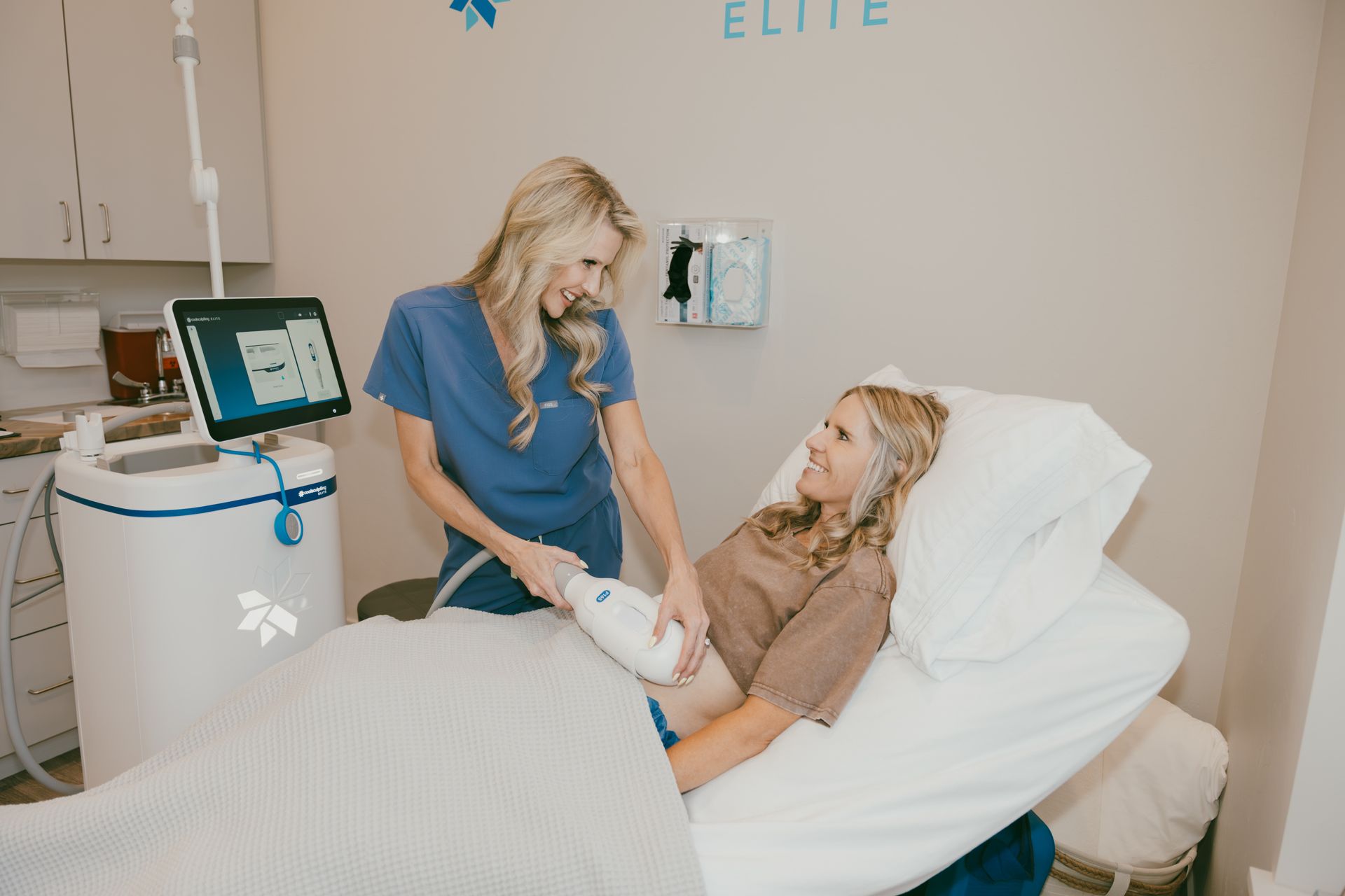 A medical professional in blue scrubs treats a patient's leg with a CoolSculpting device in a clinic. Both women smile.