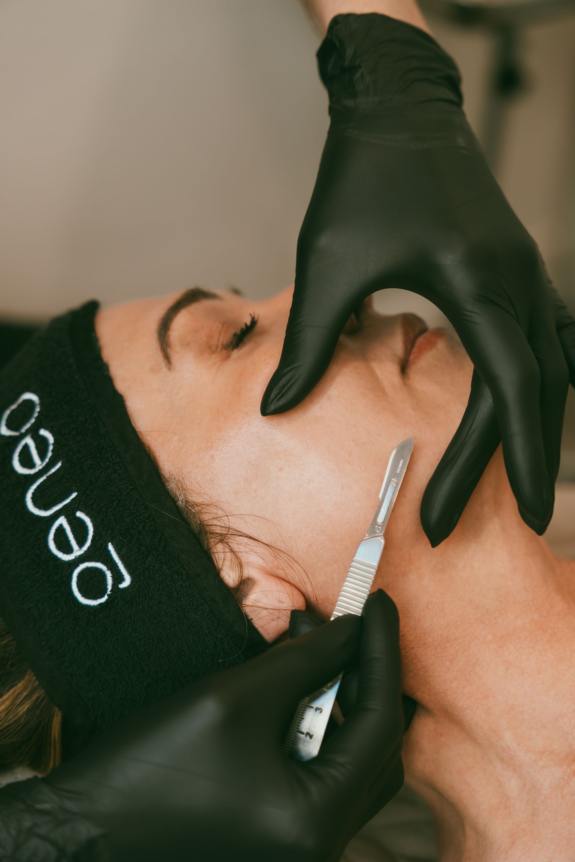 Person receiving dermaplaning treatment; gloved hands hold a scalpel to their jawline. Black headband, closed eyes.