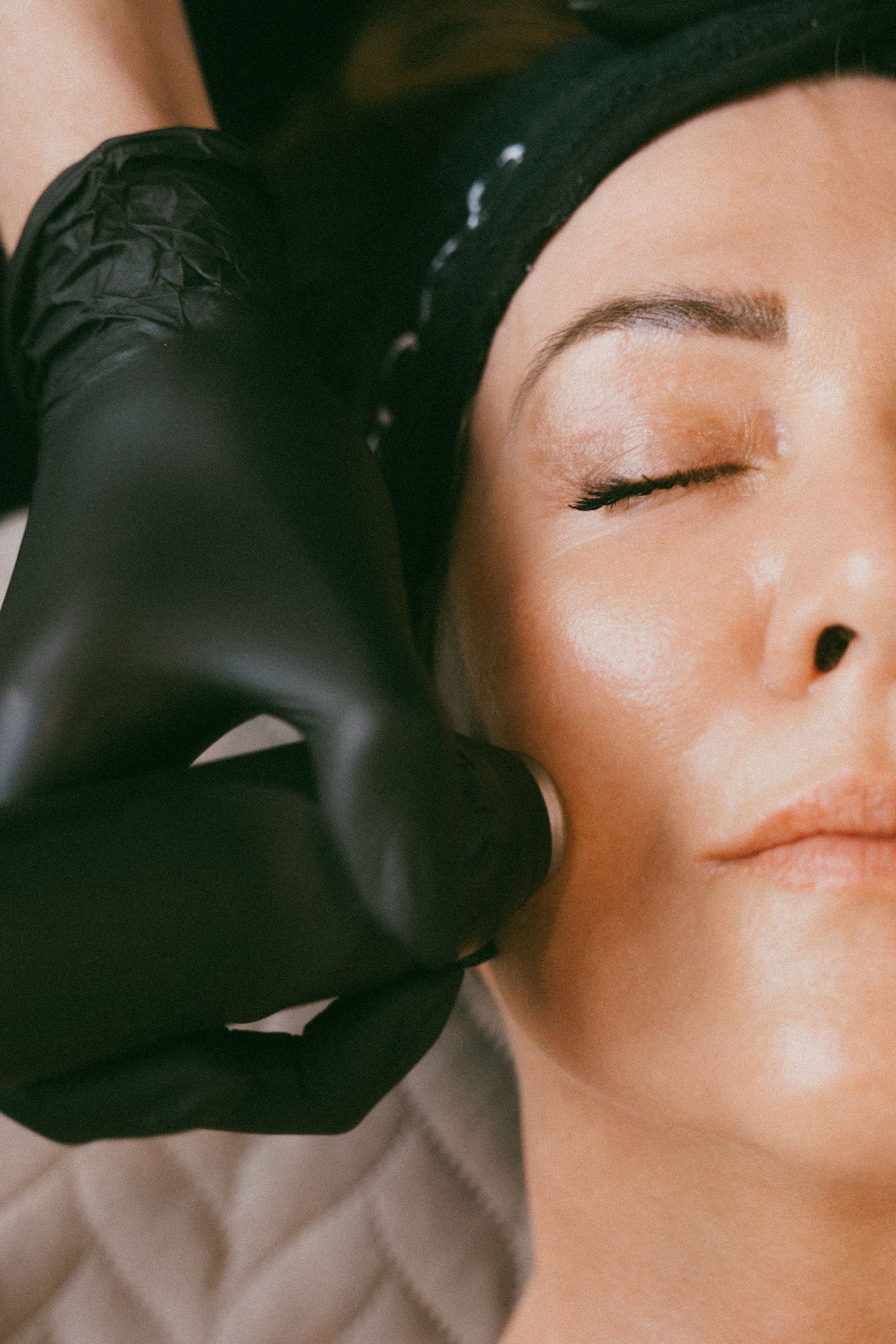 Person receiving facial treatment. A gloved hand presses against the woman's cheek; she has eyes closed.