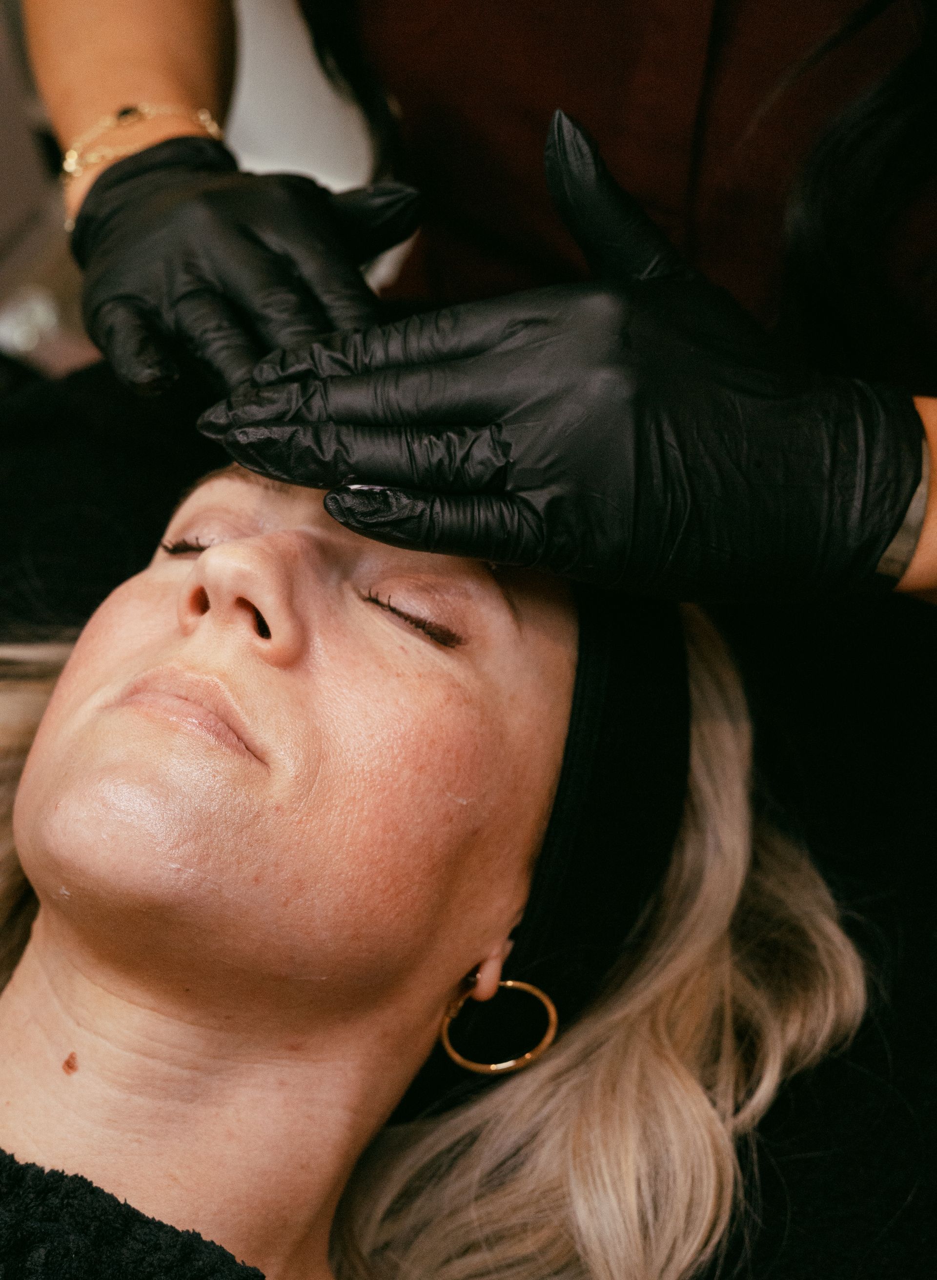 Woman receiving facial treatment, eyes closed, hands in black gloves massaging forehead.