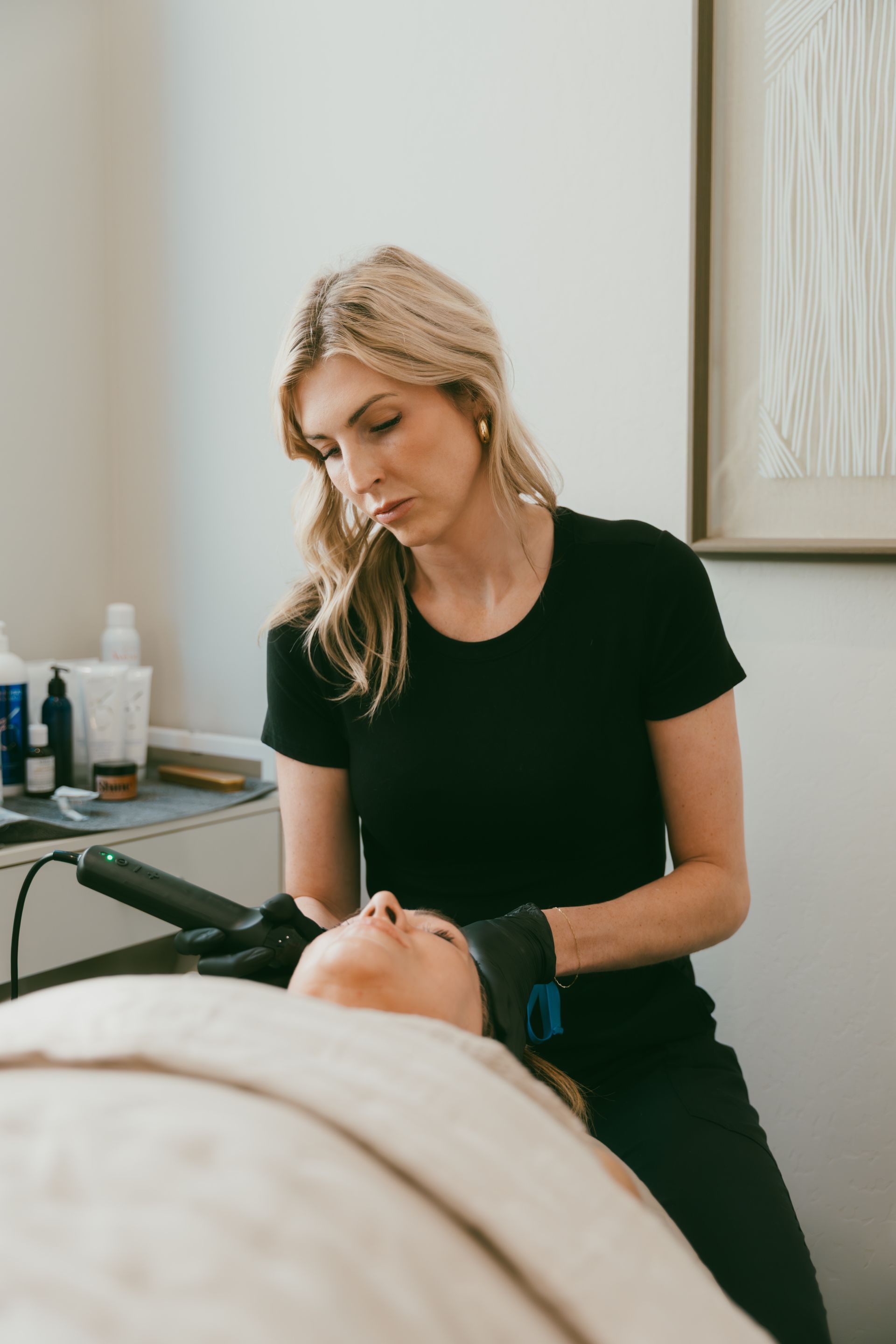 A blonde woman in black clothing performs a facial treatment on a person lying on a spa bed. She wears gloves and focuses on the client's face.