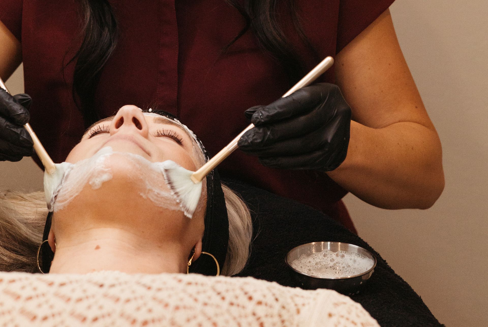 A woman receiving a facial treatment in a spa. A gloved esthetician applies a white cream to her face using small spatulas.