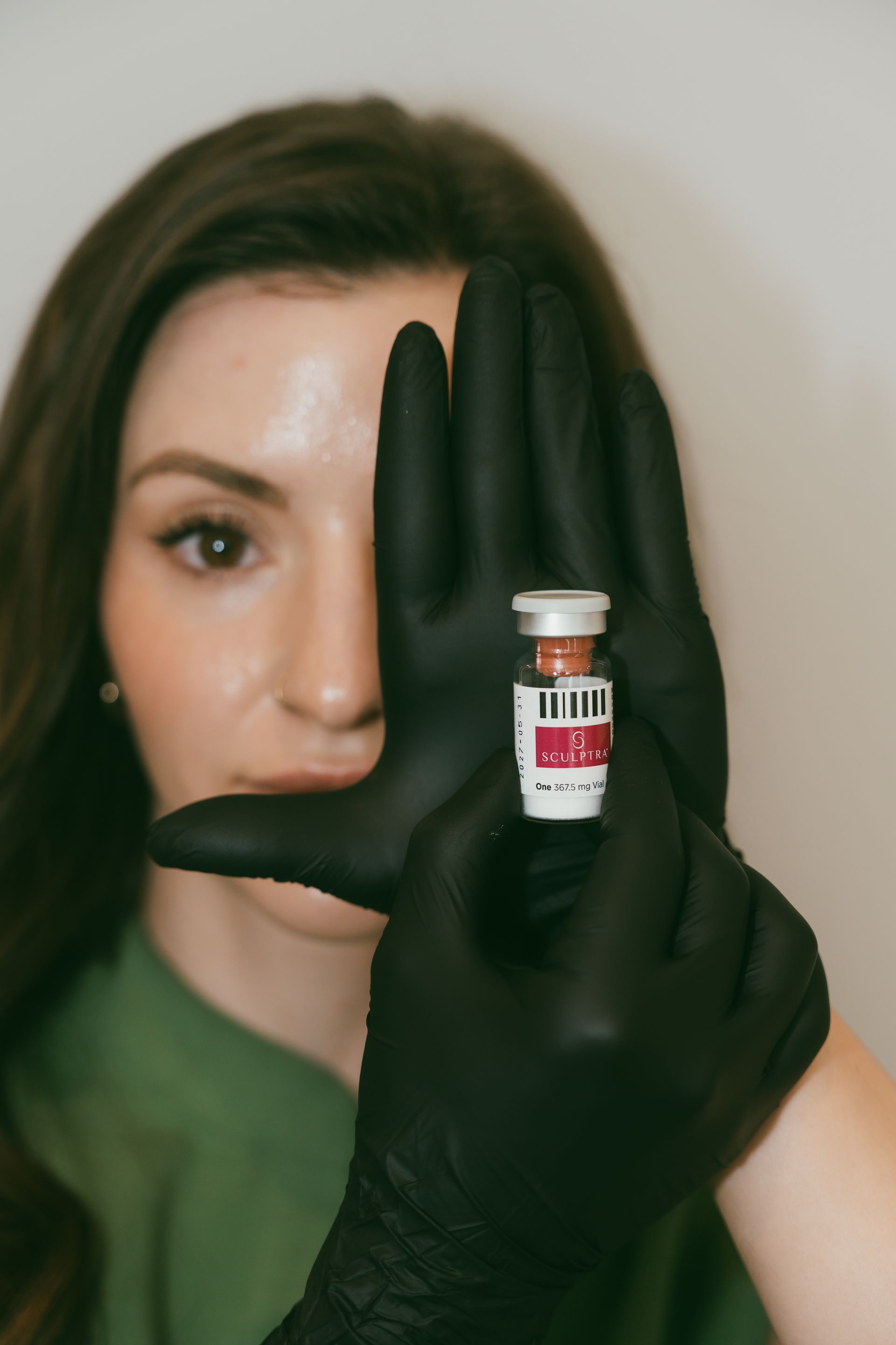 Woman holding a vial with gloved hands in front of her face. She is wearing a green top.