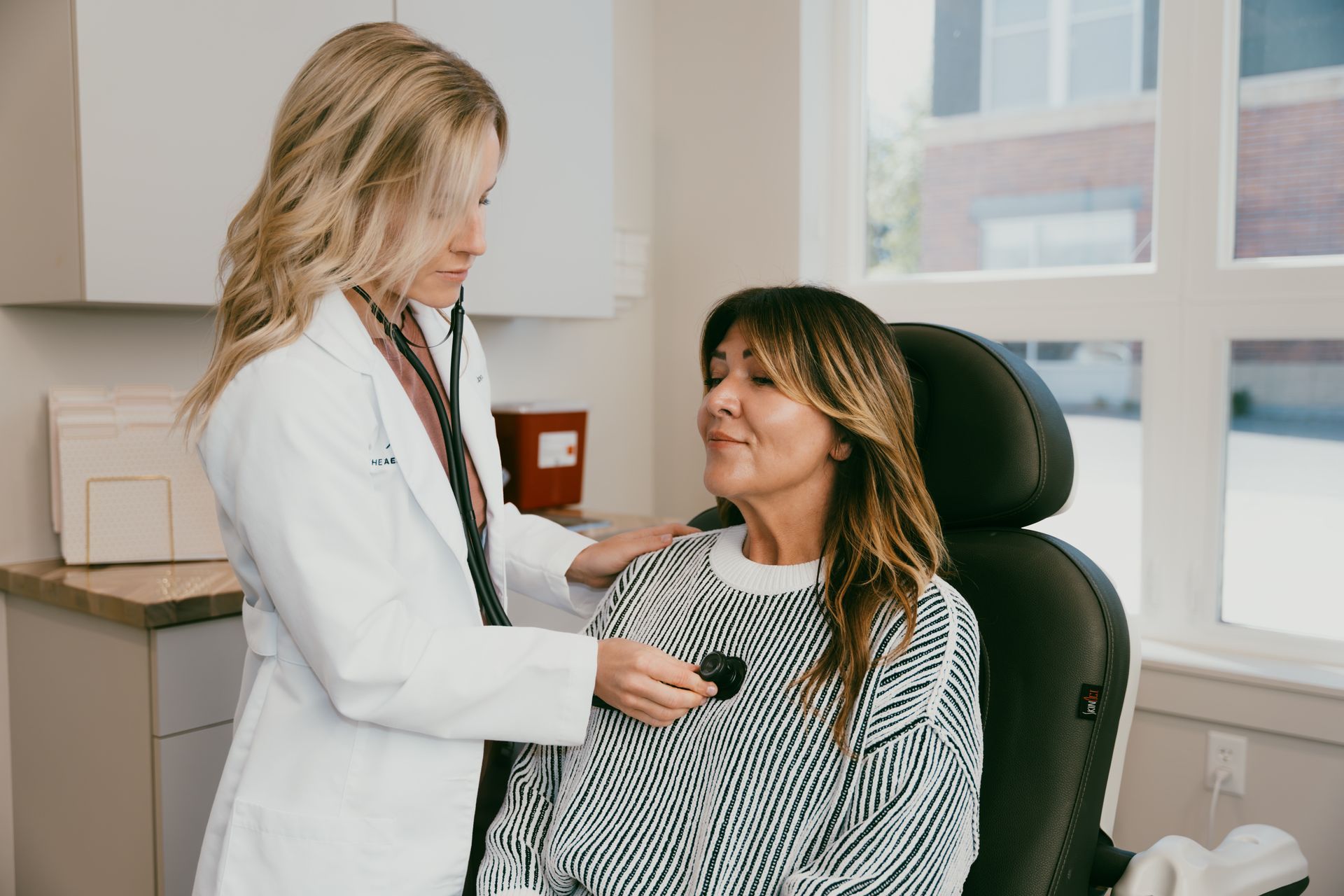 A doctor listens to a patient's chest with a stethoscope in a medical office. The patient smiles.