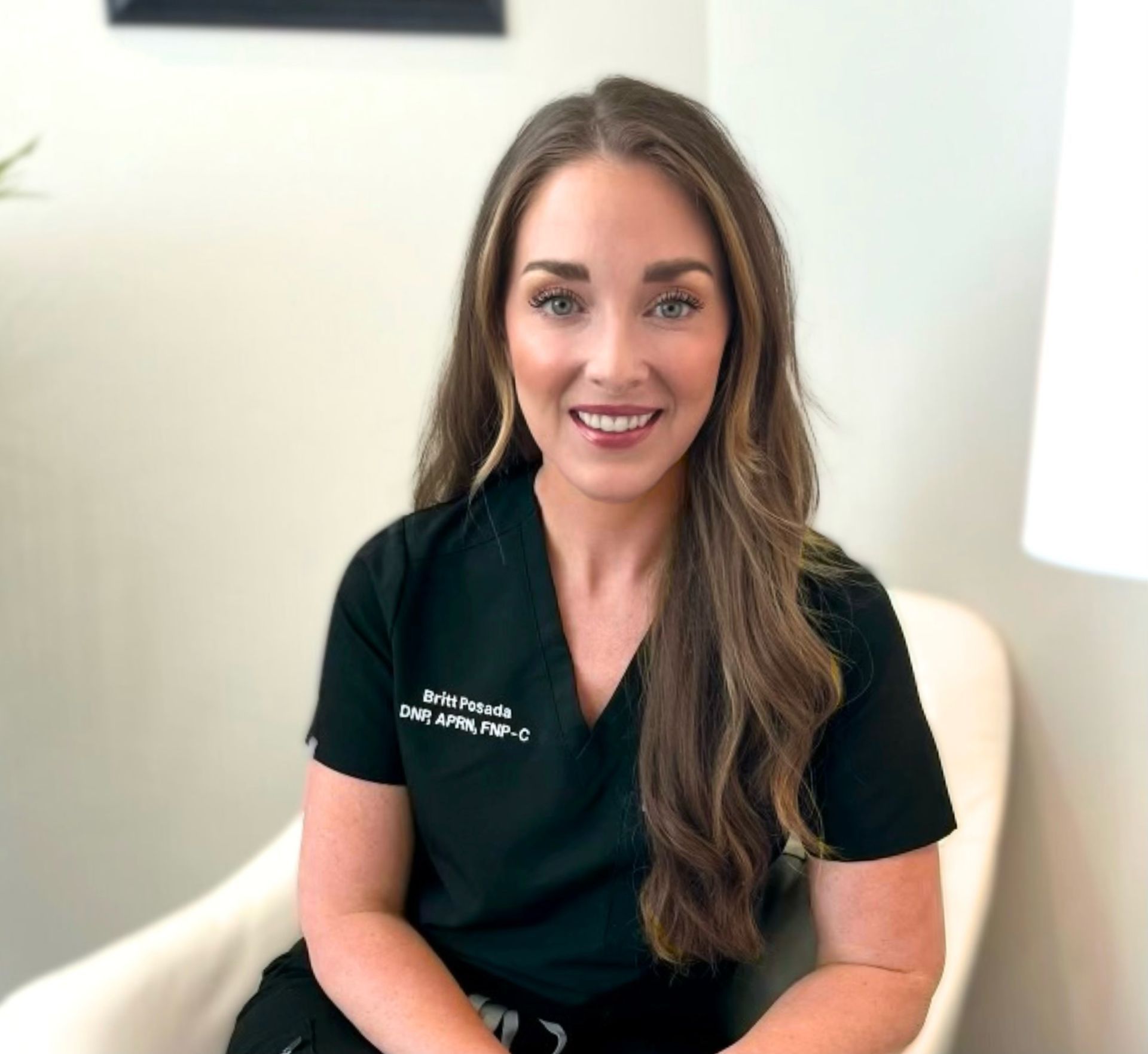 A woman in a black scrub top is sitting in a chair and smiling in The Aesthetic Firm