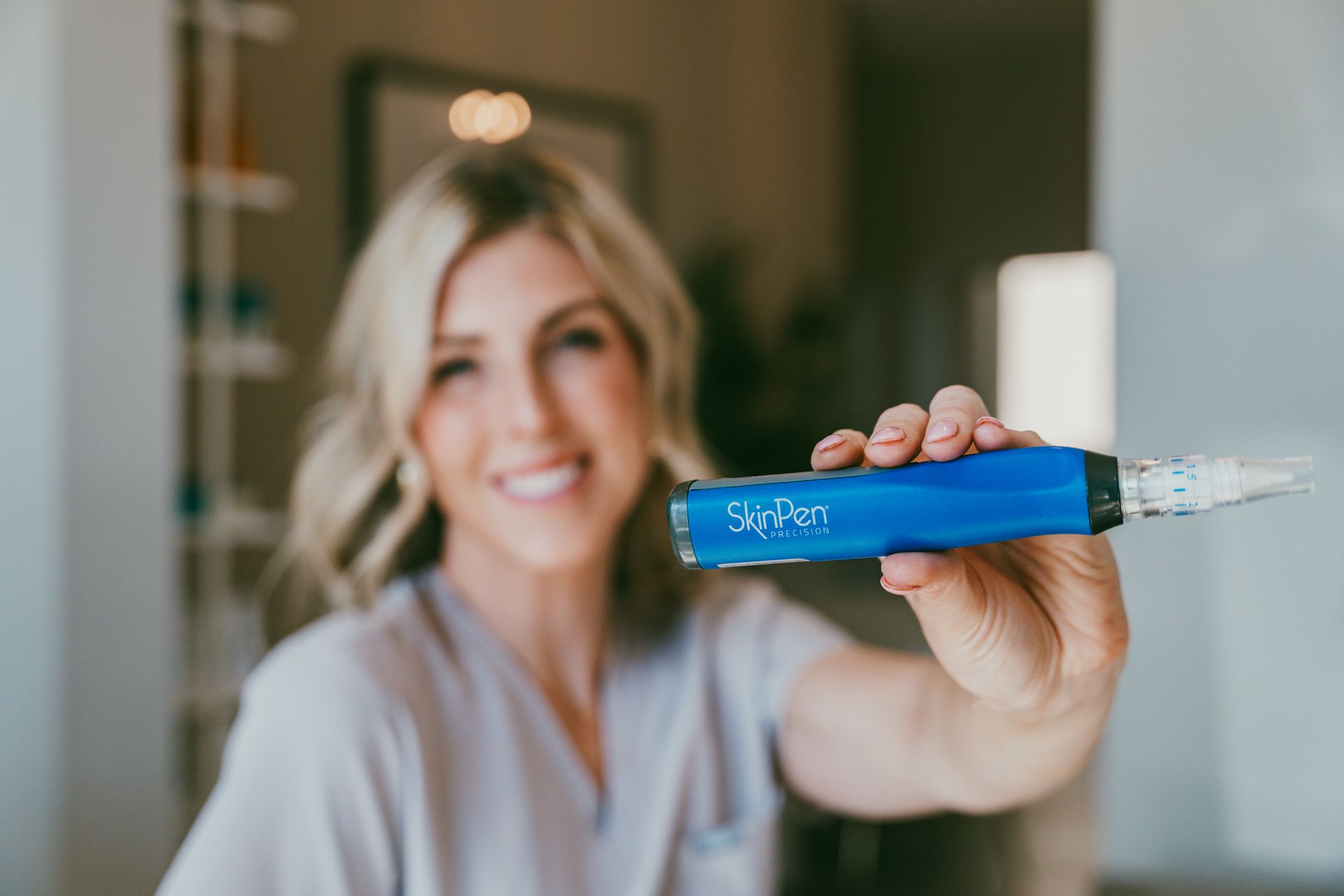 Blonde woman in scrubs smiling, holding a blue Skin Pen device, indoors.