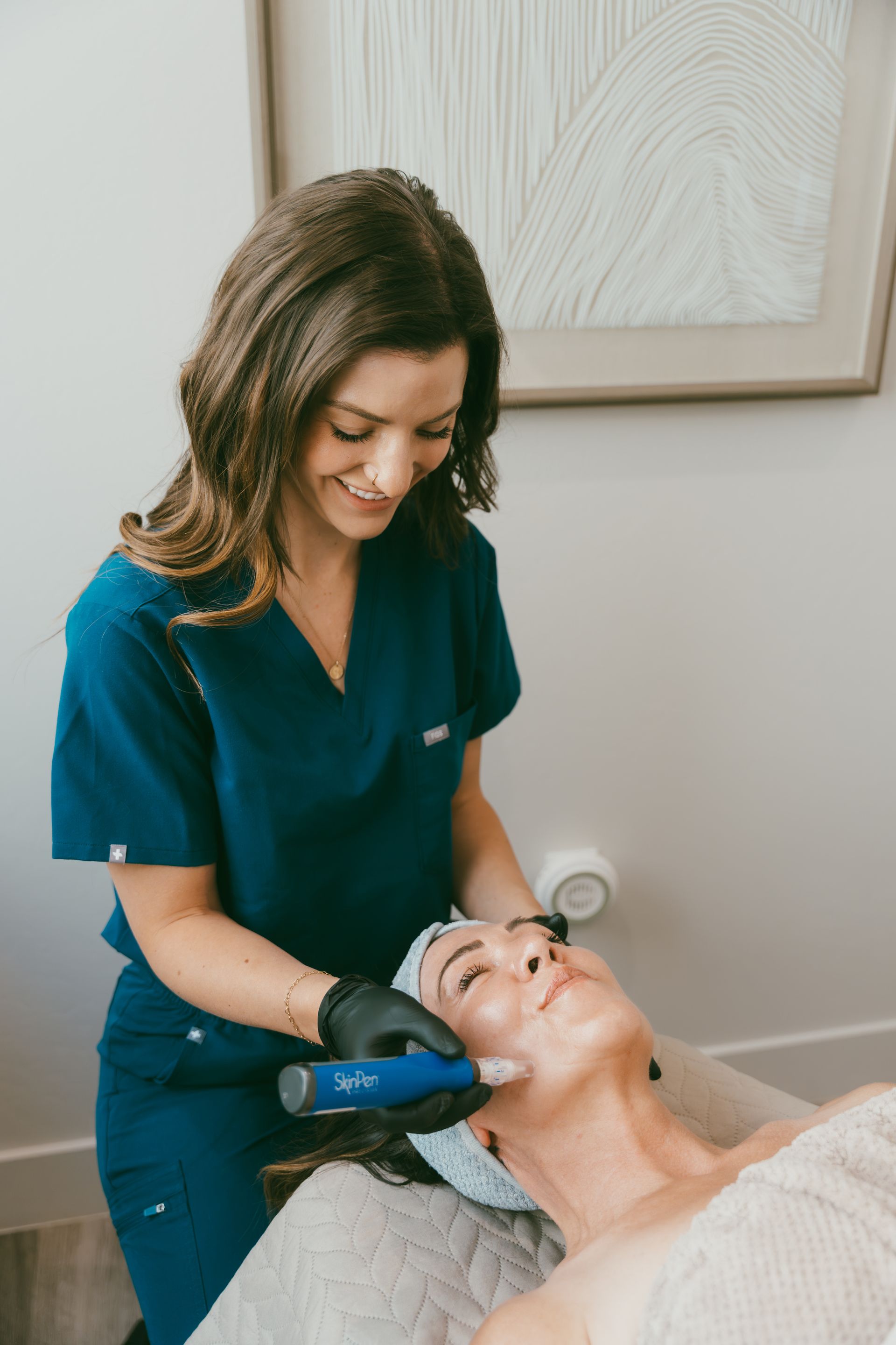 Woman in blue scrubs performing a skin treatment on another woman lying down; both smiling in a well-lit medical setting.