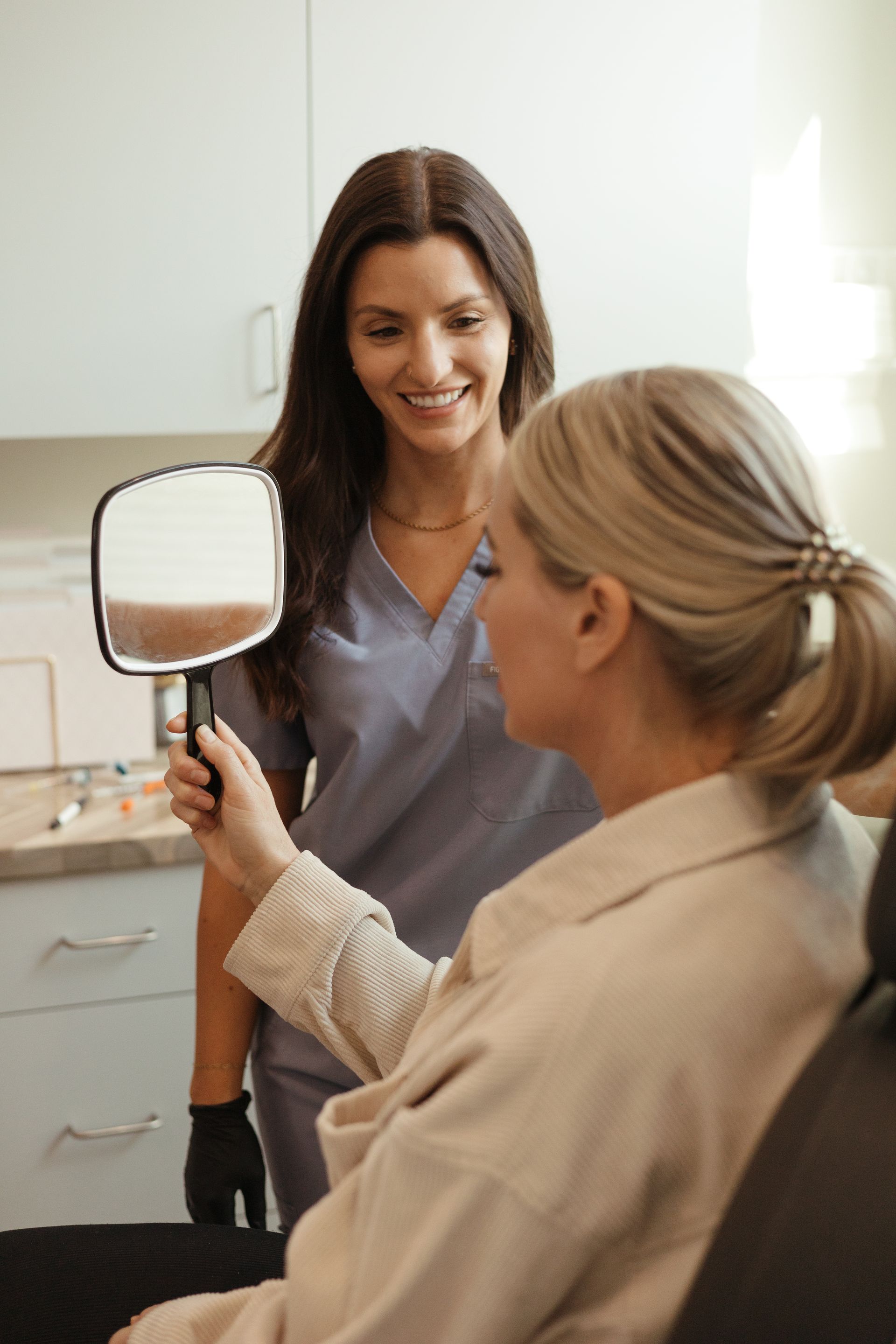 Woman looking in mirror held by a smiling person in scrubs; clinic setting.