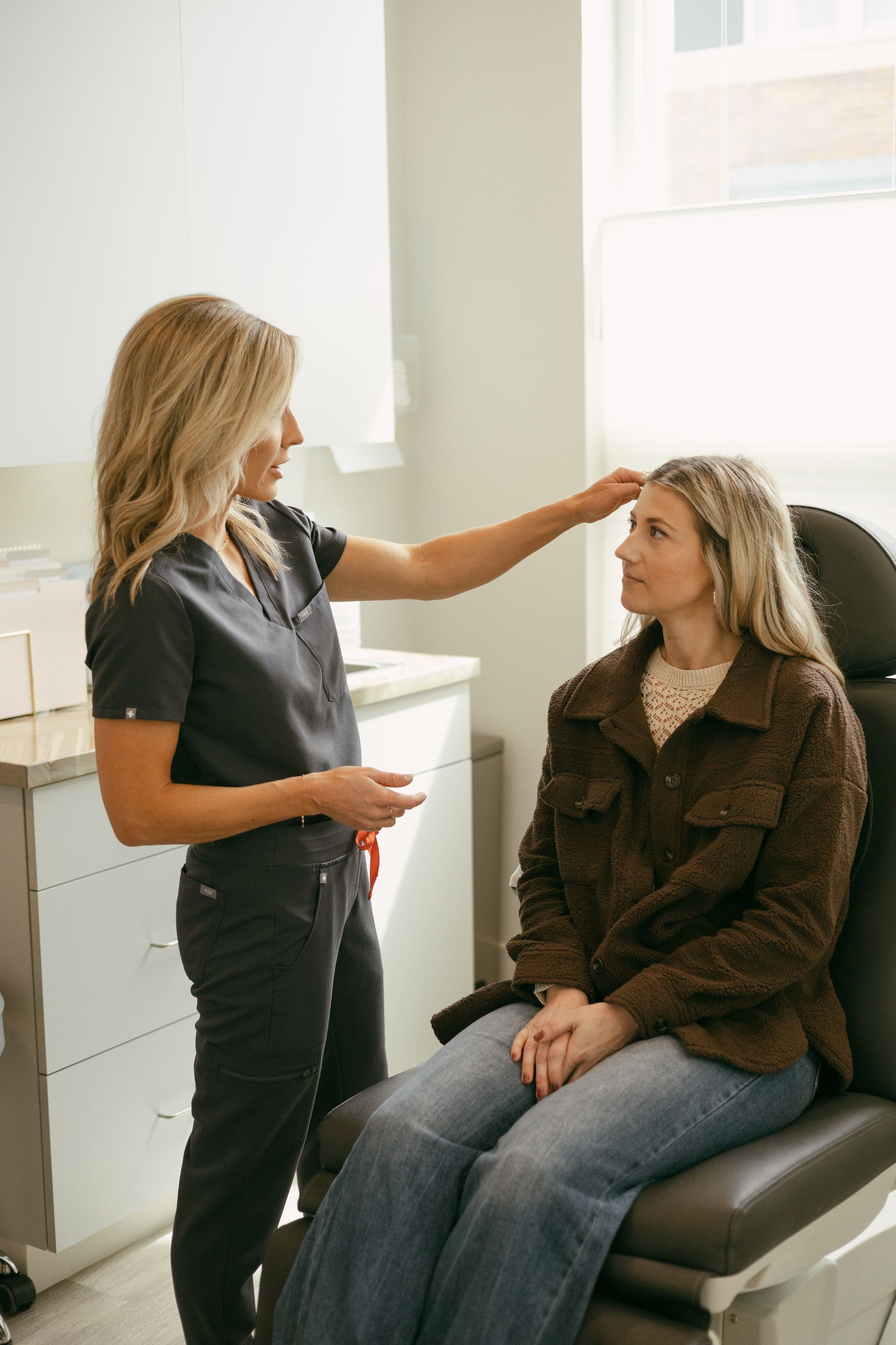 Woman examining another woman's forehead in medical setting; one woman seated, wearing a jacket; the other in scrubs.