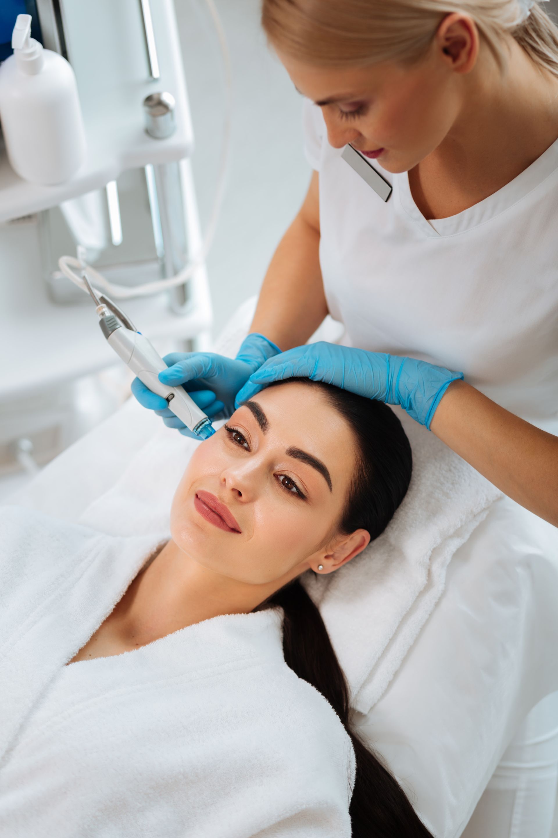 Woman receiving facial treatment in a clinic. Aesthetitican in blue gloves uses a device on her face.