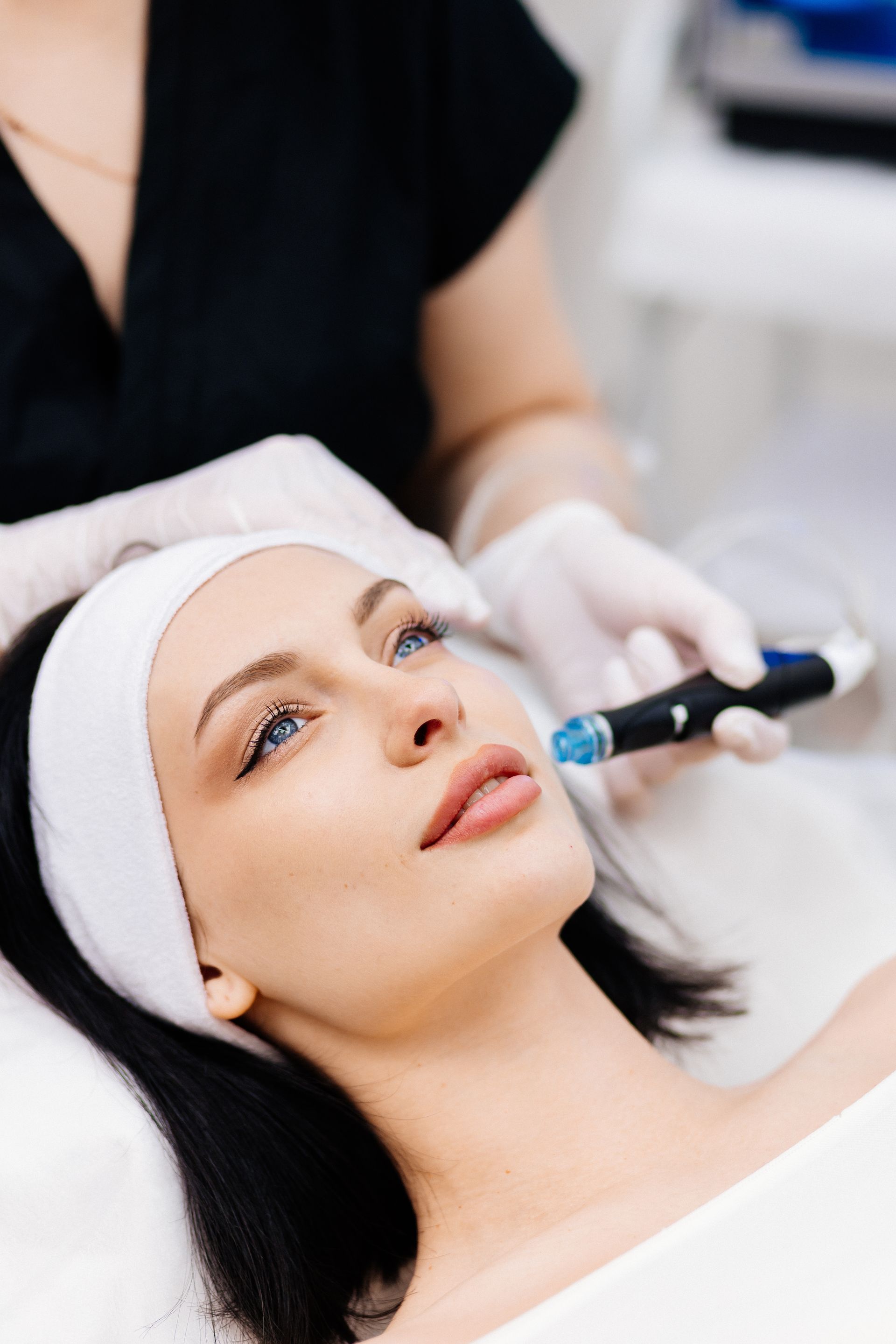 Woman receiving facial treatment; cosmetician holding device near face, wearing gloves.