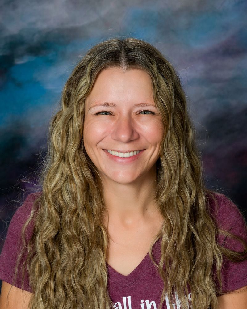 A woman with long blonde hair is smiling and wearing a purple shirt.