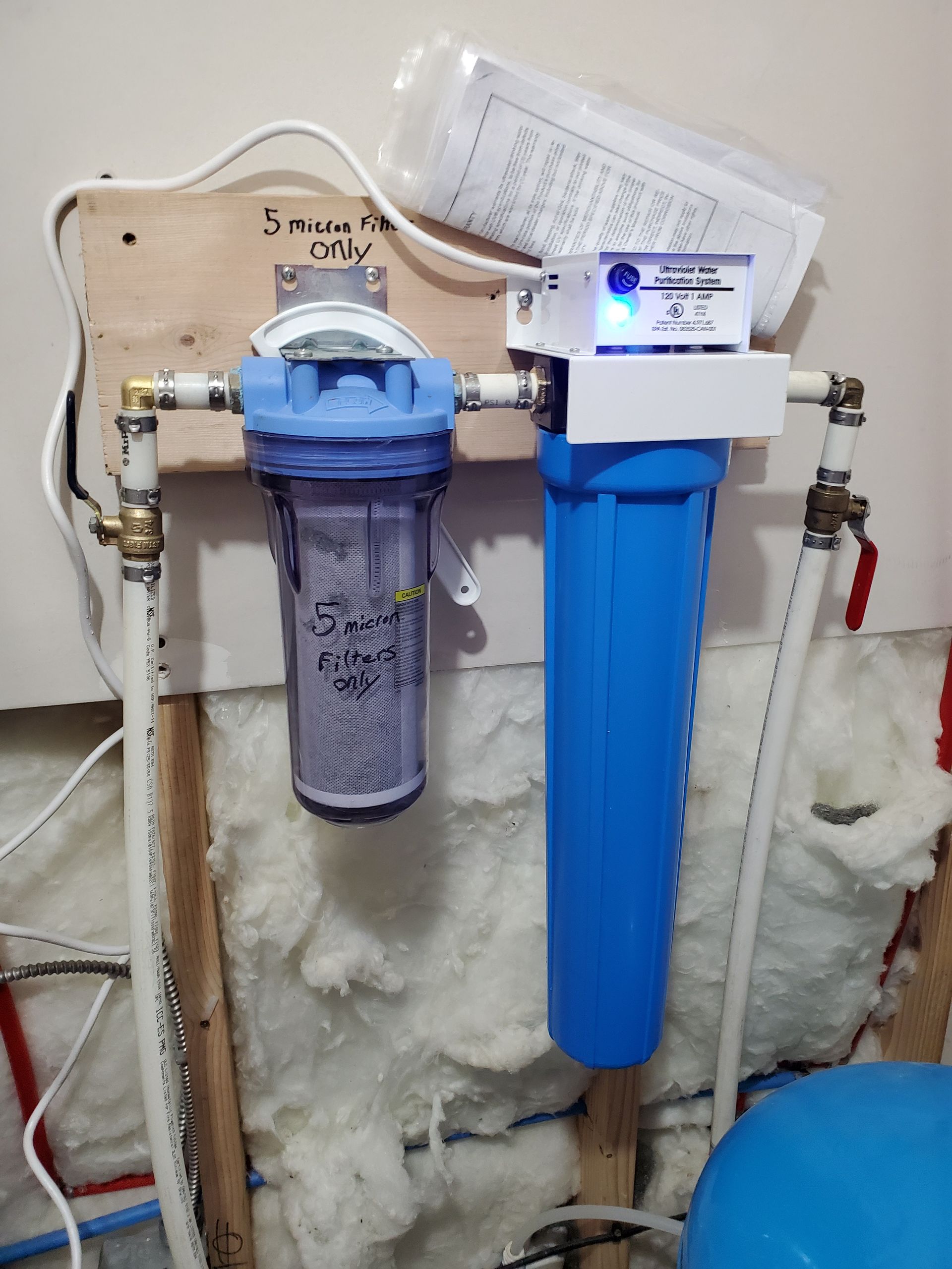 Water filtration system with blue and clear filter housings, mounted on a wooden board. Plumbing connections visible.