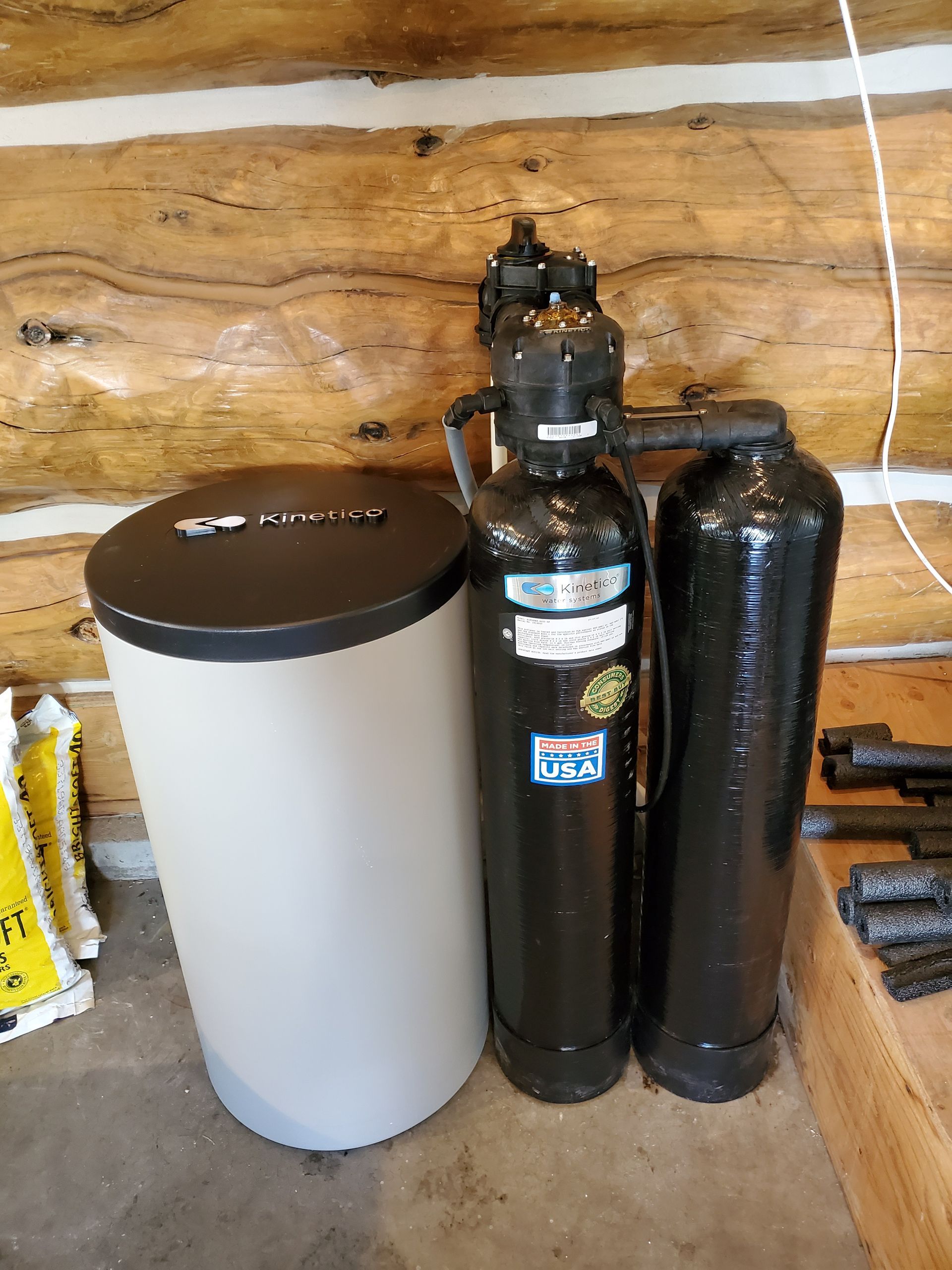 Water softener system with a tall, light-colored tank and two black tanks against a wood-paneled wall.