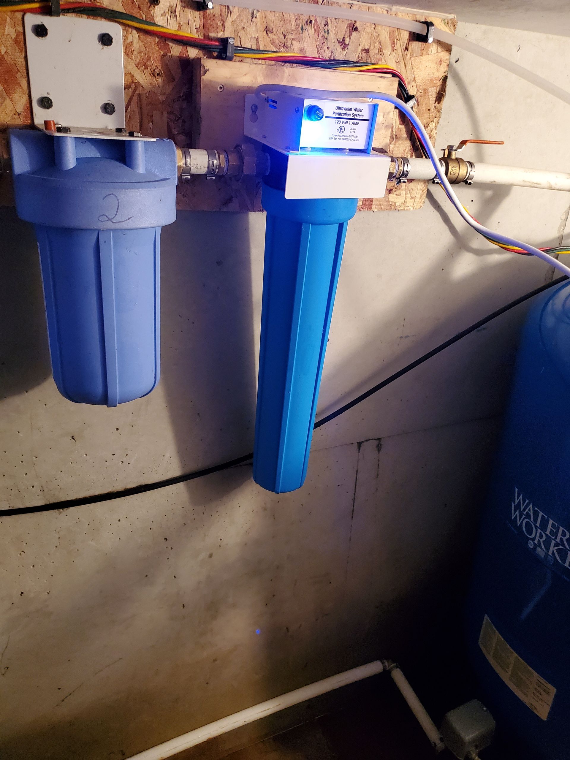 Two blue water filter housings on a wooden wall, with a control unit and a pressure tank visible.