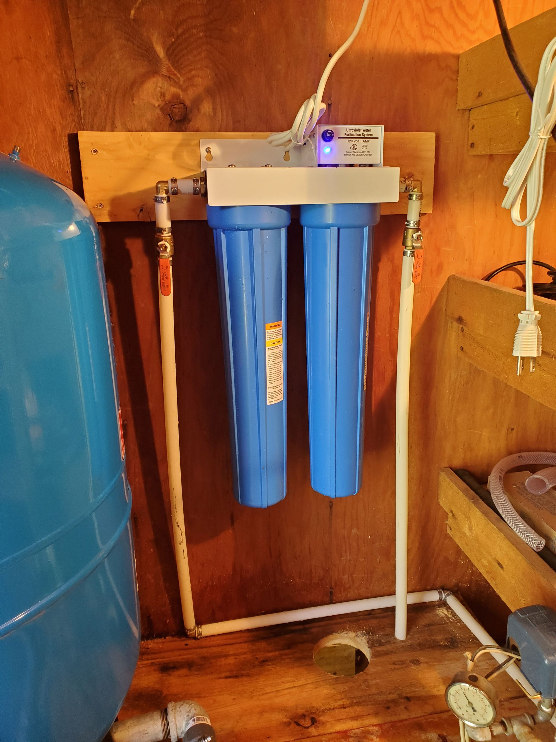 Water filtration system with two blue filter housings, mounted on a wooden board. Piping and a pressure tank are visible.