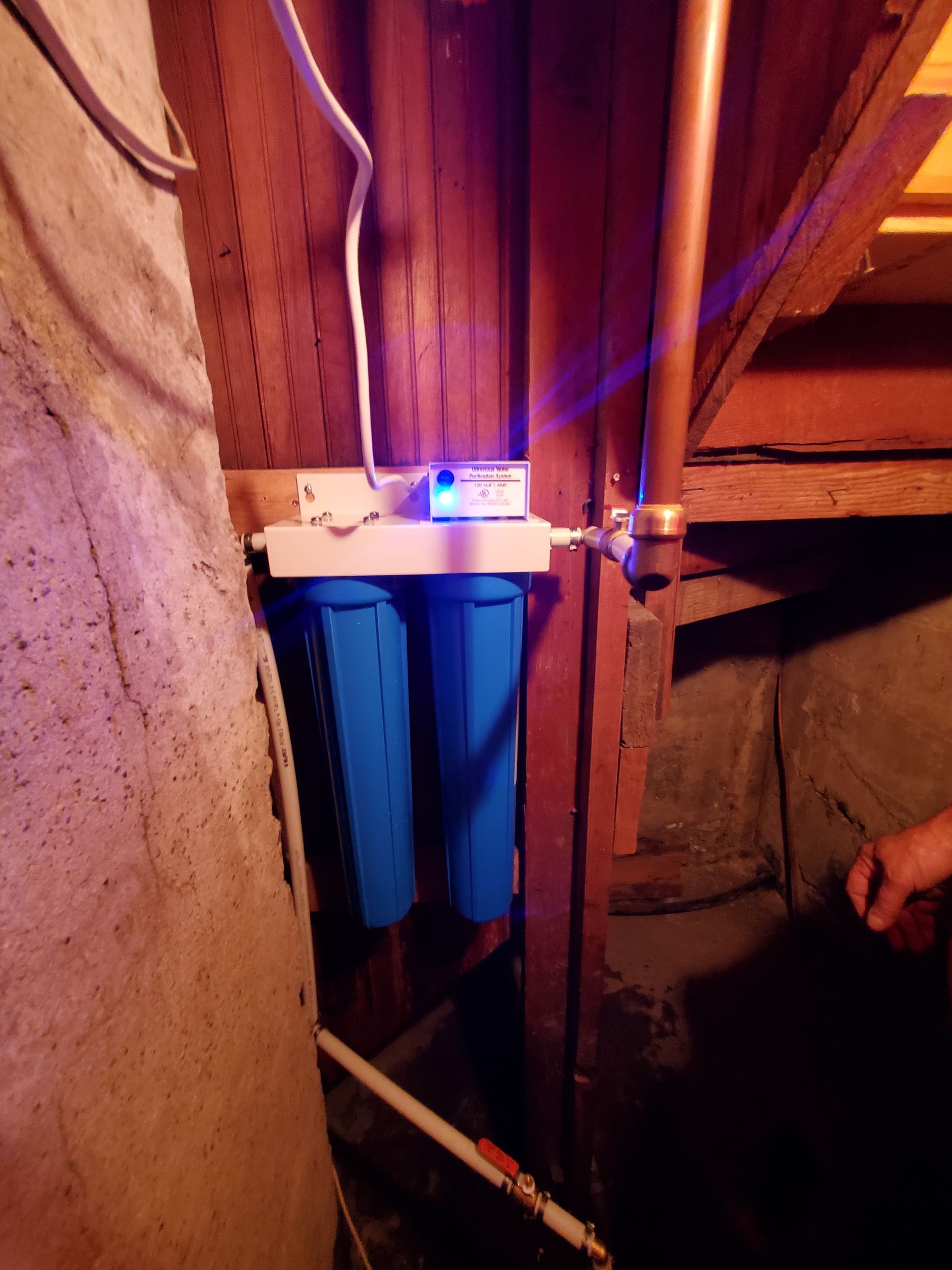 Blue water filter system with copper pipe, mounted on wood in a dimly lit basement.