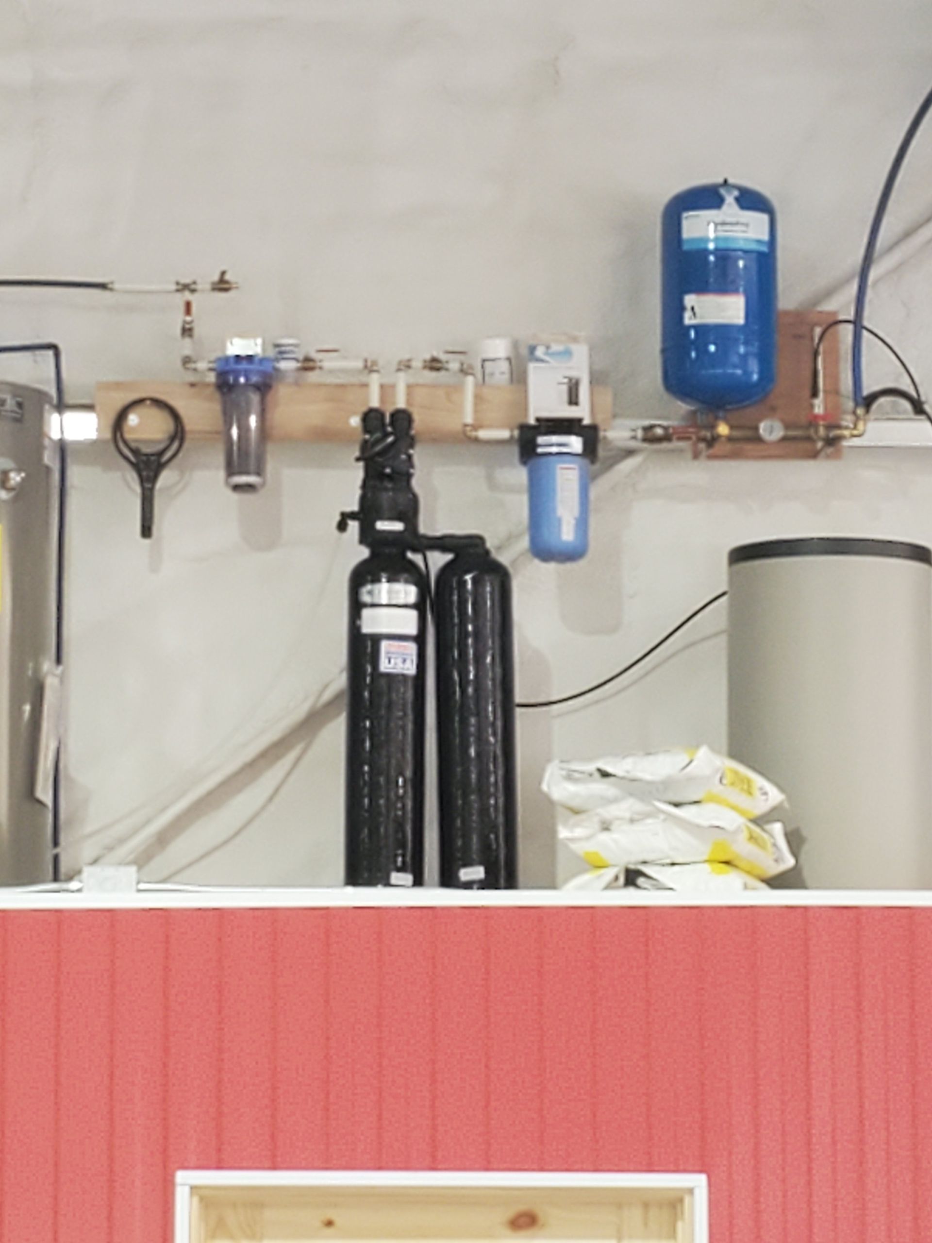 Water filtration system with tanks, filters, and pressure tank mounted on a wall above red paneling.