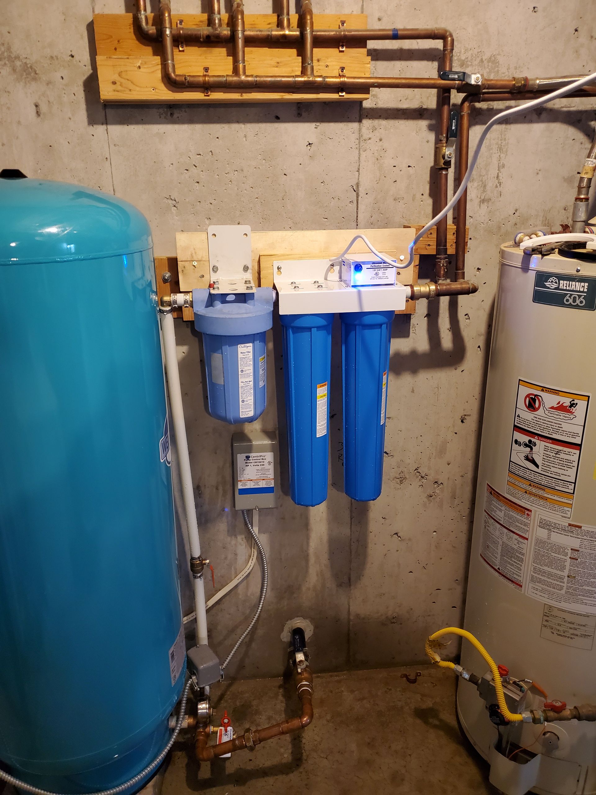 Water filtration system with blue tanks, pipes, and a water heater against a concrete wall.