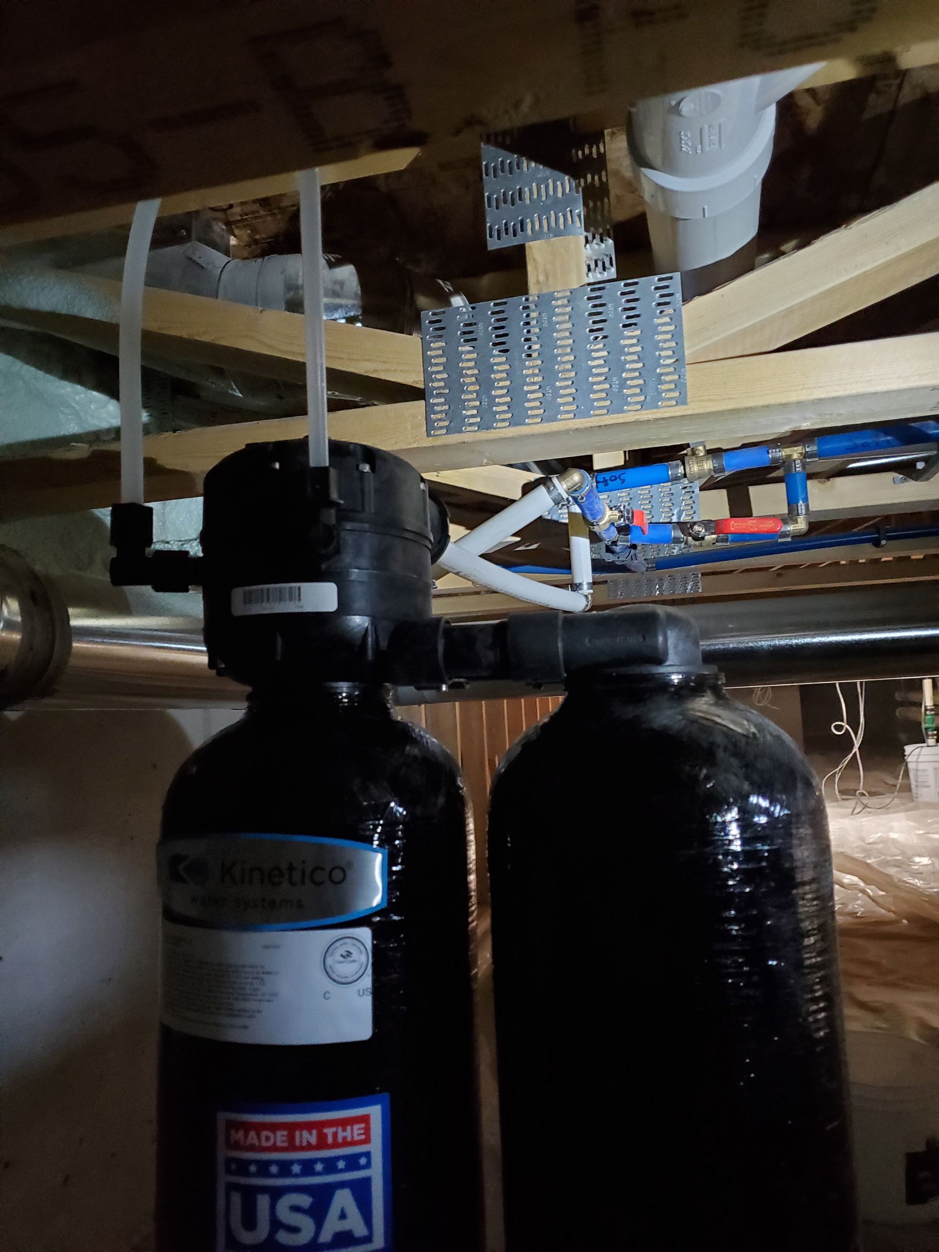 Two black water filter tanks under wooden ceiling joists, with white pipes and blue PEX plumbing visible.