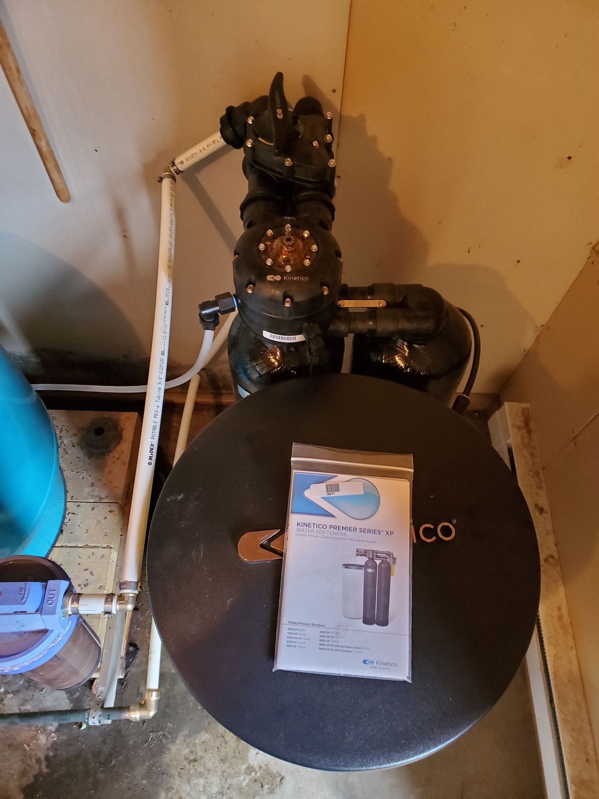 Water filtration system with black tank, control head, and bag of replacement parts. Beige walls.