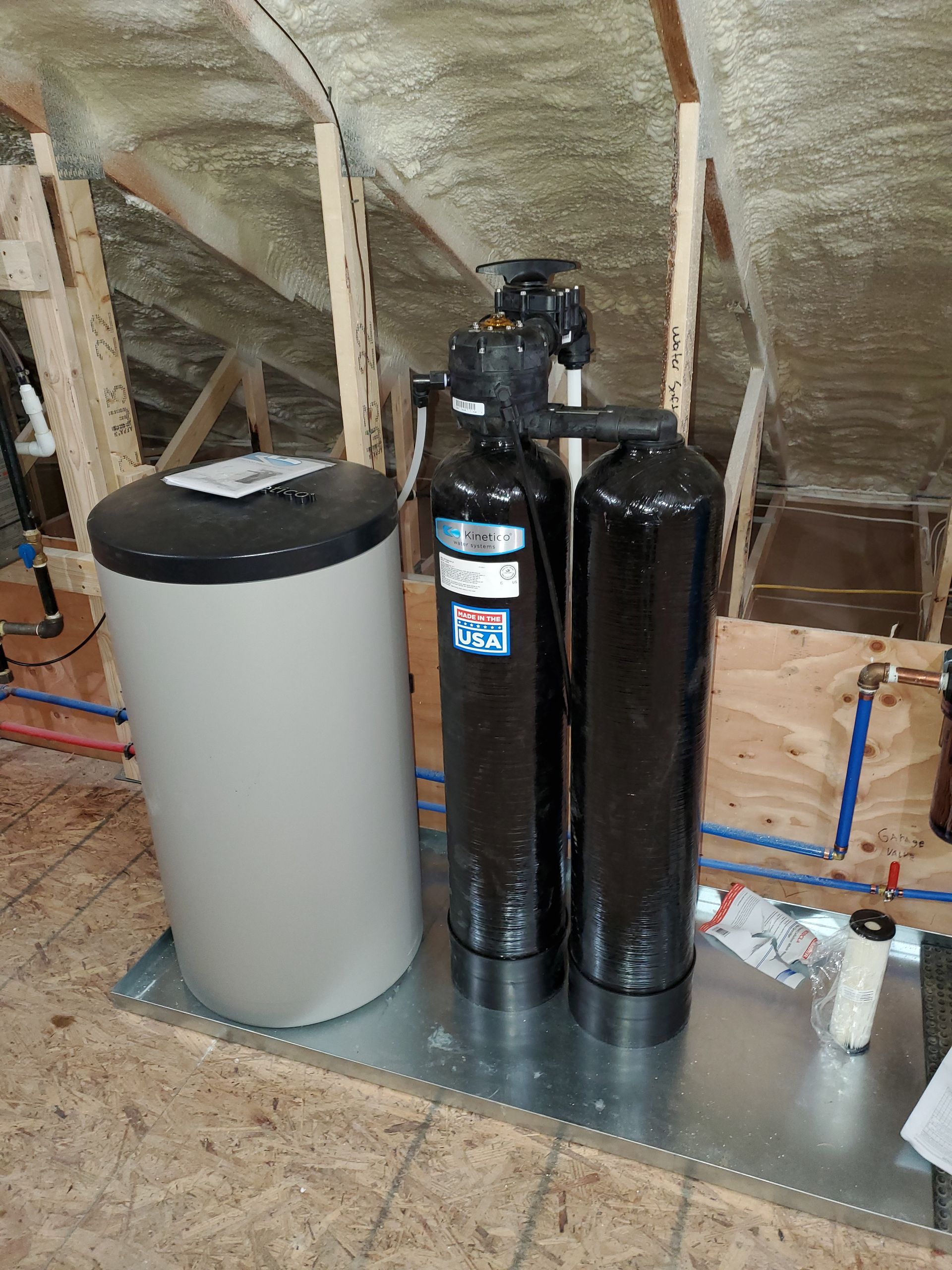 Water softener system in attic: tank, filter tanks, and control valve. Installed on metal platform with wood framing.