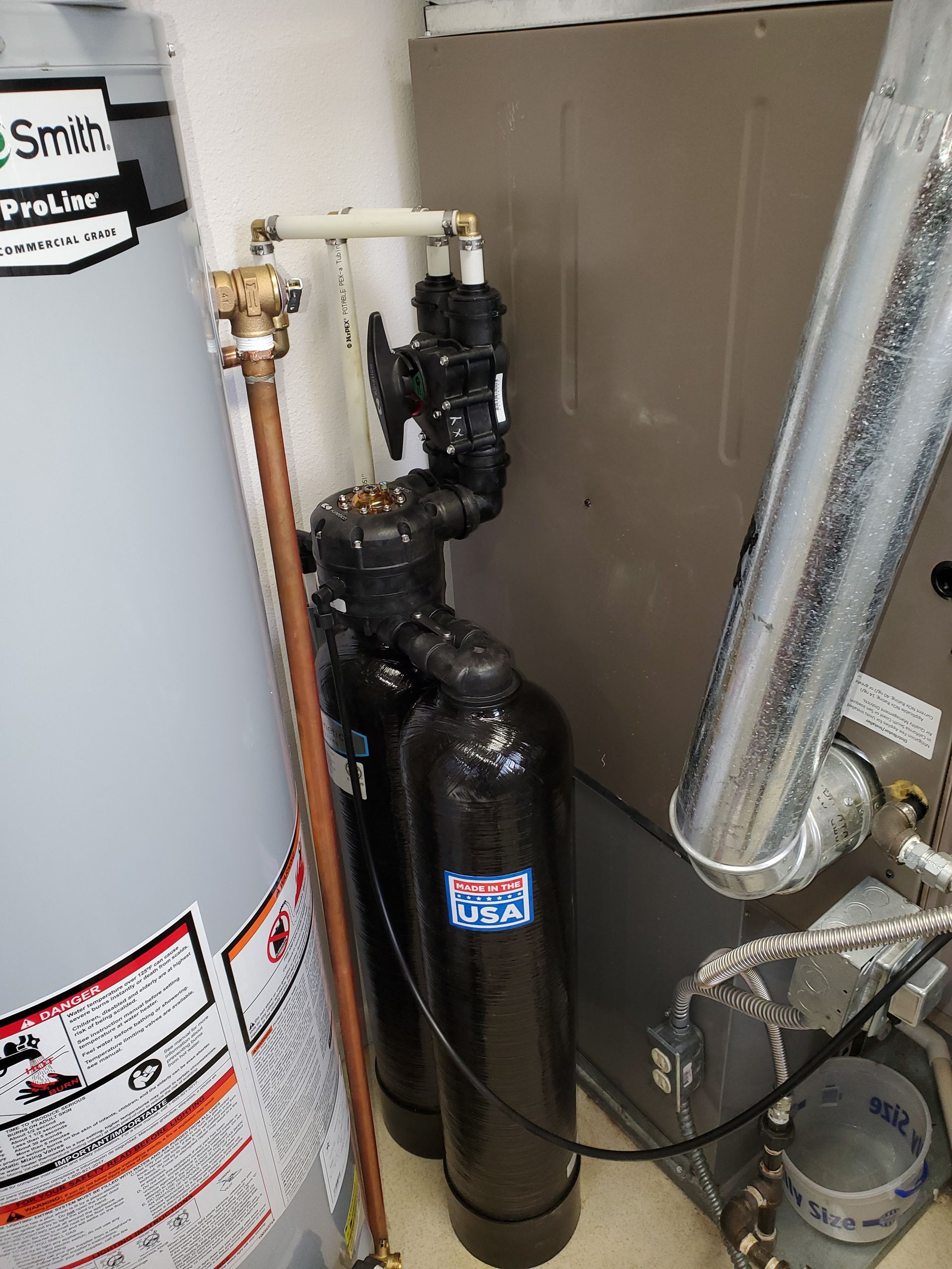 Water softener system beside a hot water heater and furnace. Black tanks, copper and white pipes.