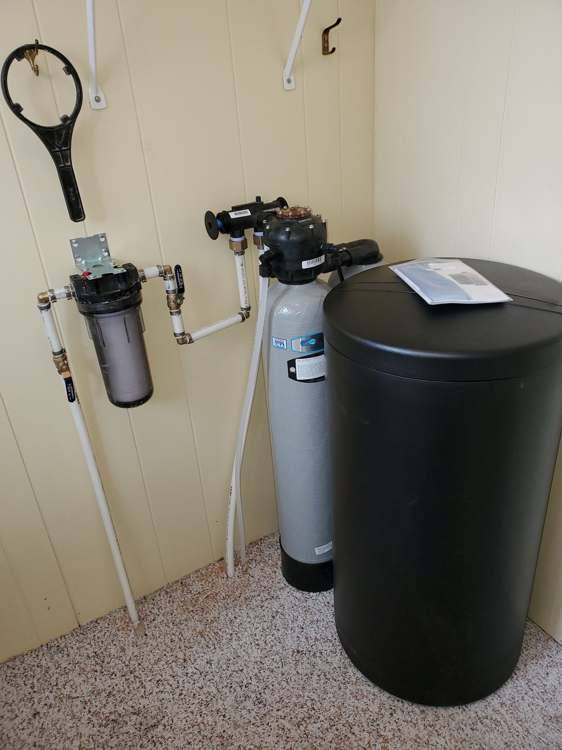 Water filtration system in a utility room; black and gray tanks, white pipes on a tan wall.