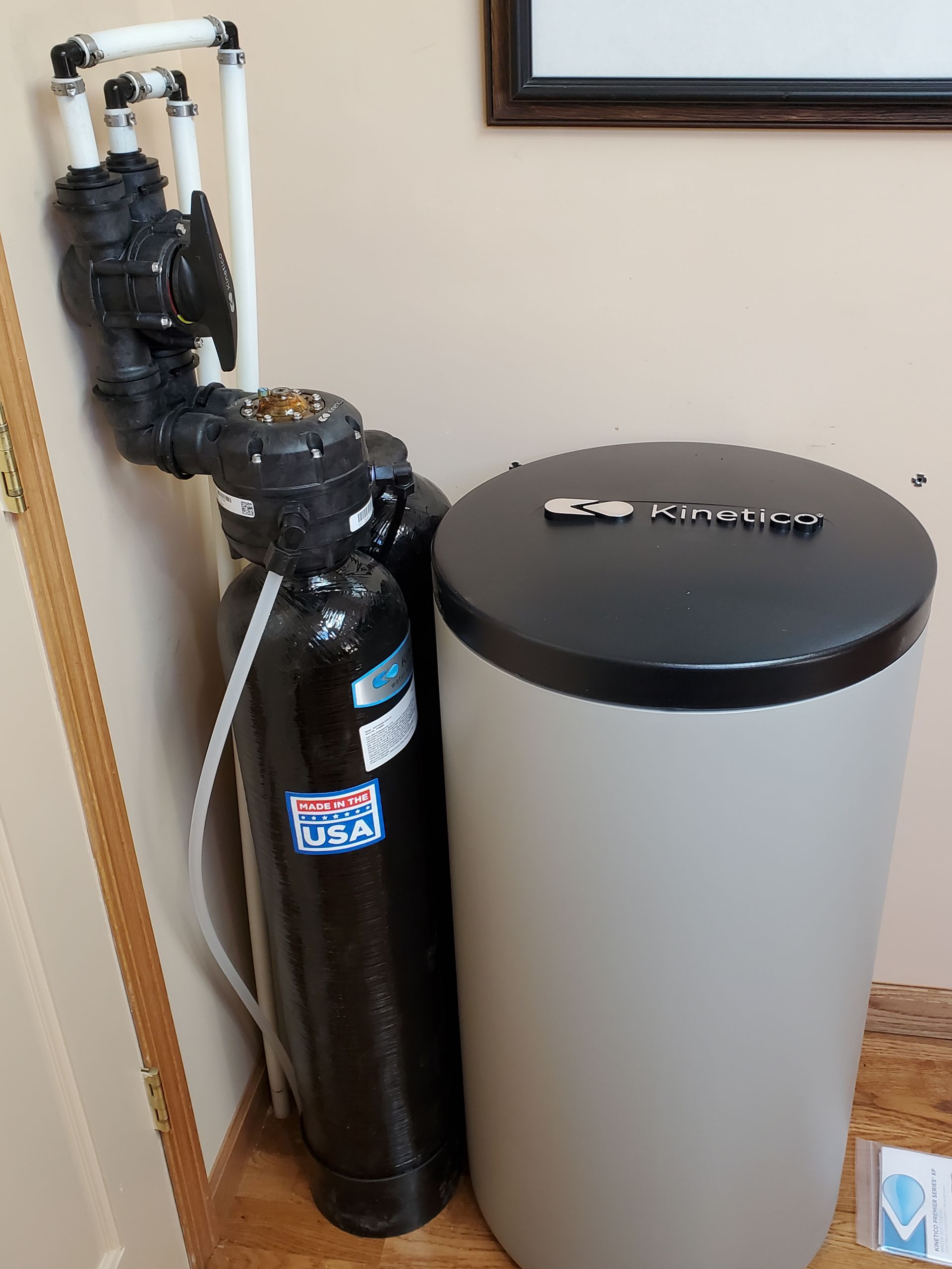 Water softener system with black tank, valve, and salt storage container against a beige wall.