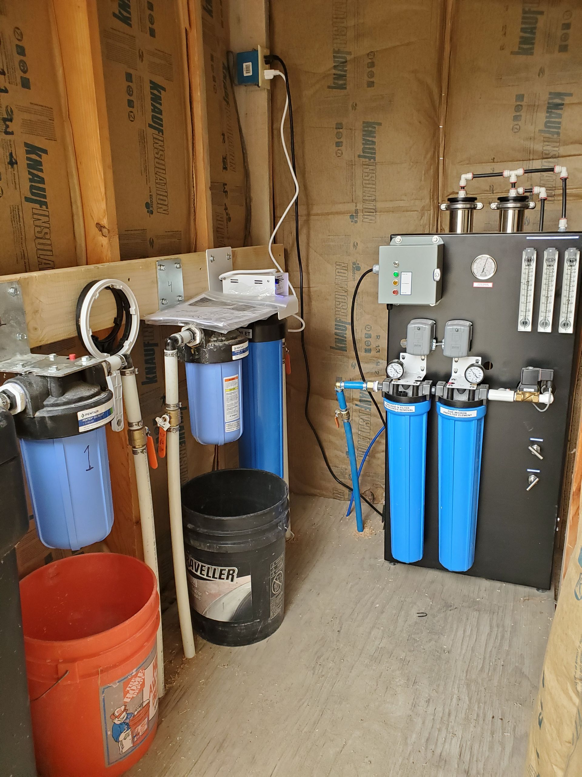 Water filtration system in a utility room with blue and black filters, pipes, and electrical components.