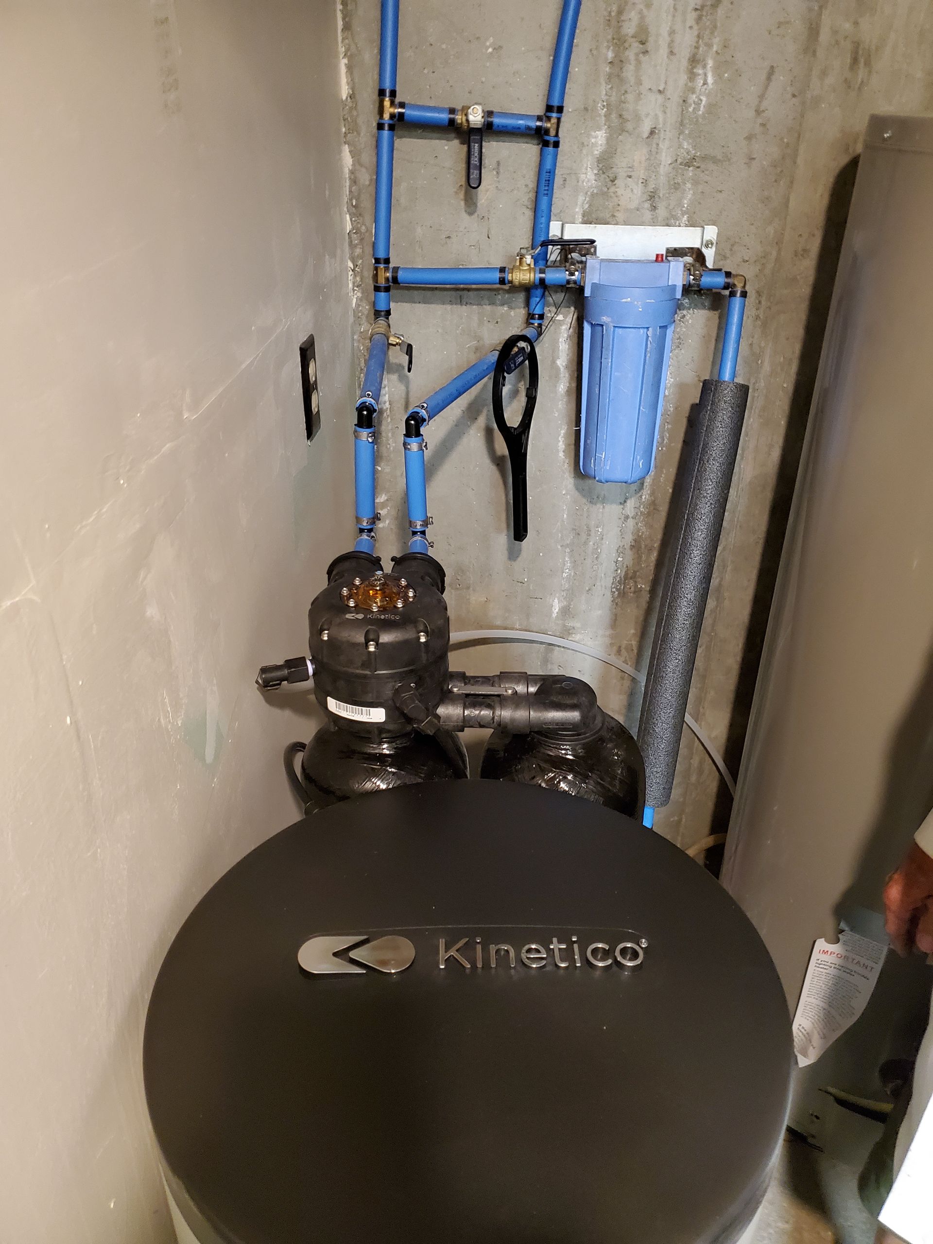 A water softener system with a black tank, blue pipes, and a filter in a basement.