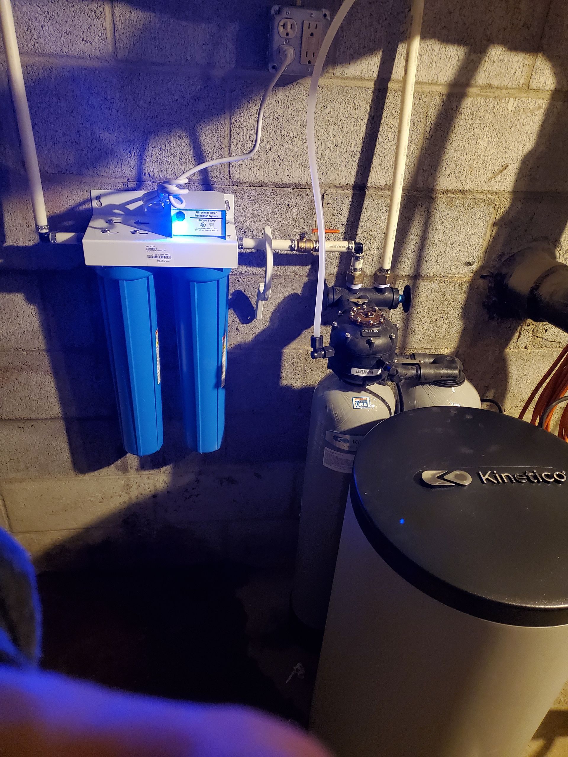 Water filtration system with blue filters, digital display, and pipes against a cinder block wall.