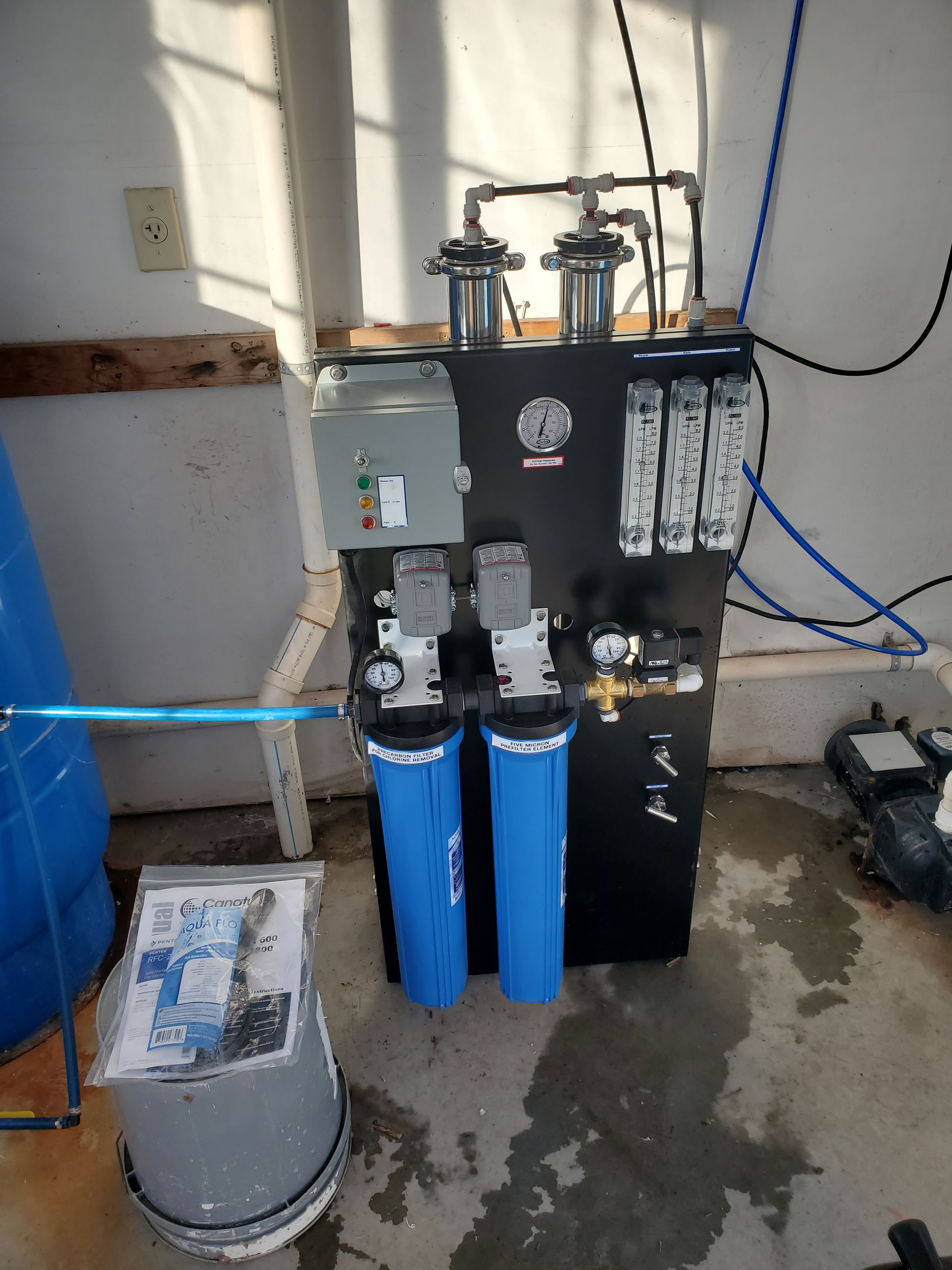 A water filtration system in a garage setting, featuring blue filter housings, black control panel, and various pipes.