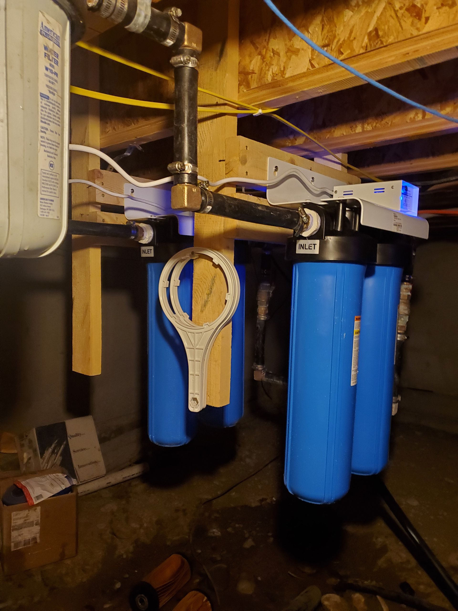 Blue water filter system installed in a basement, pipes and wood supports visible.