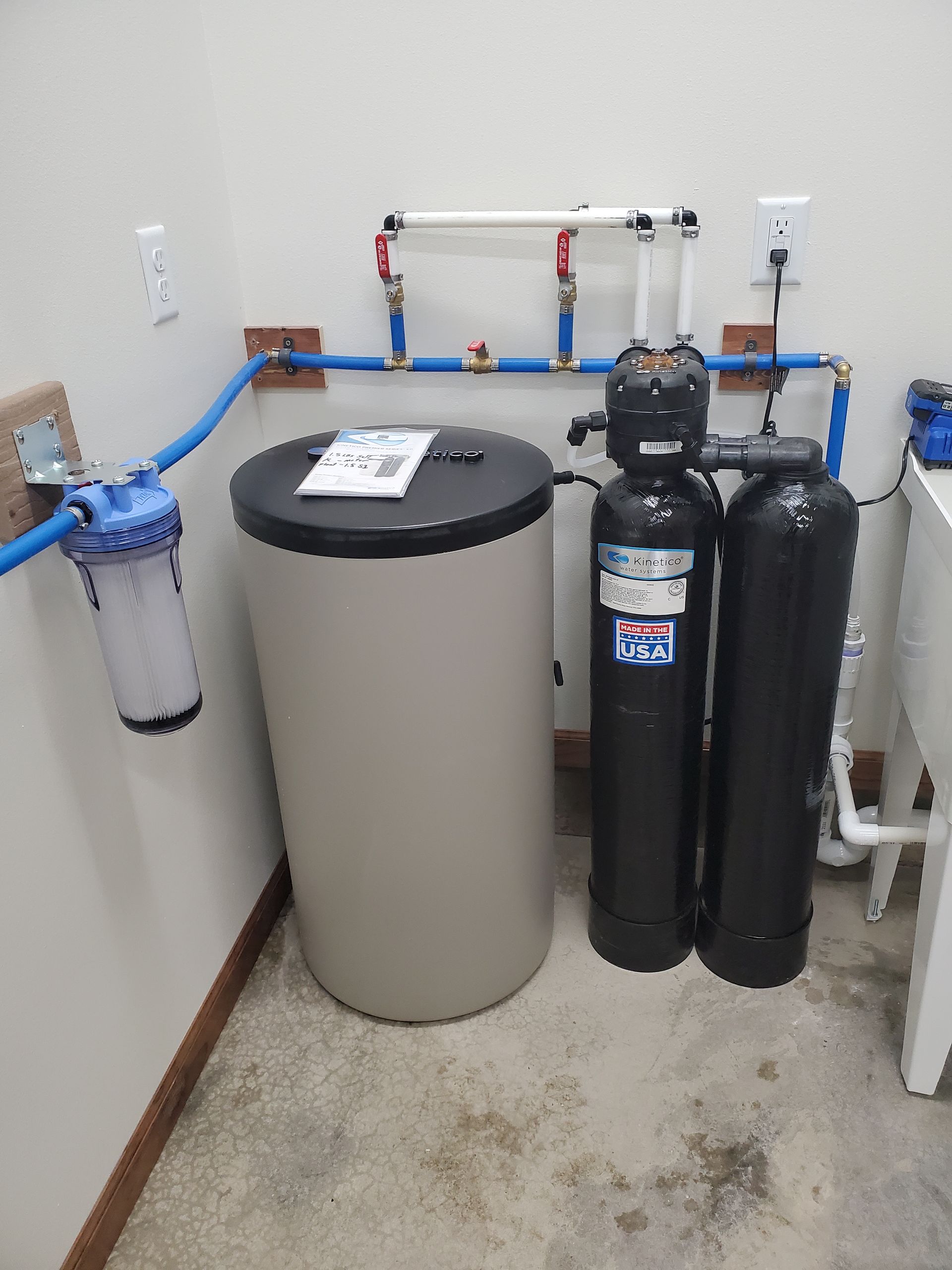 Water filtration system in a utility room; features large tank, two black cylinders, and a blue pre-filter.