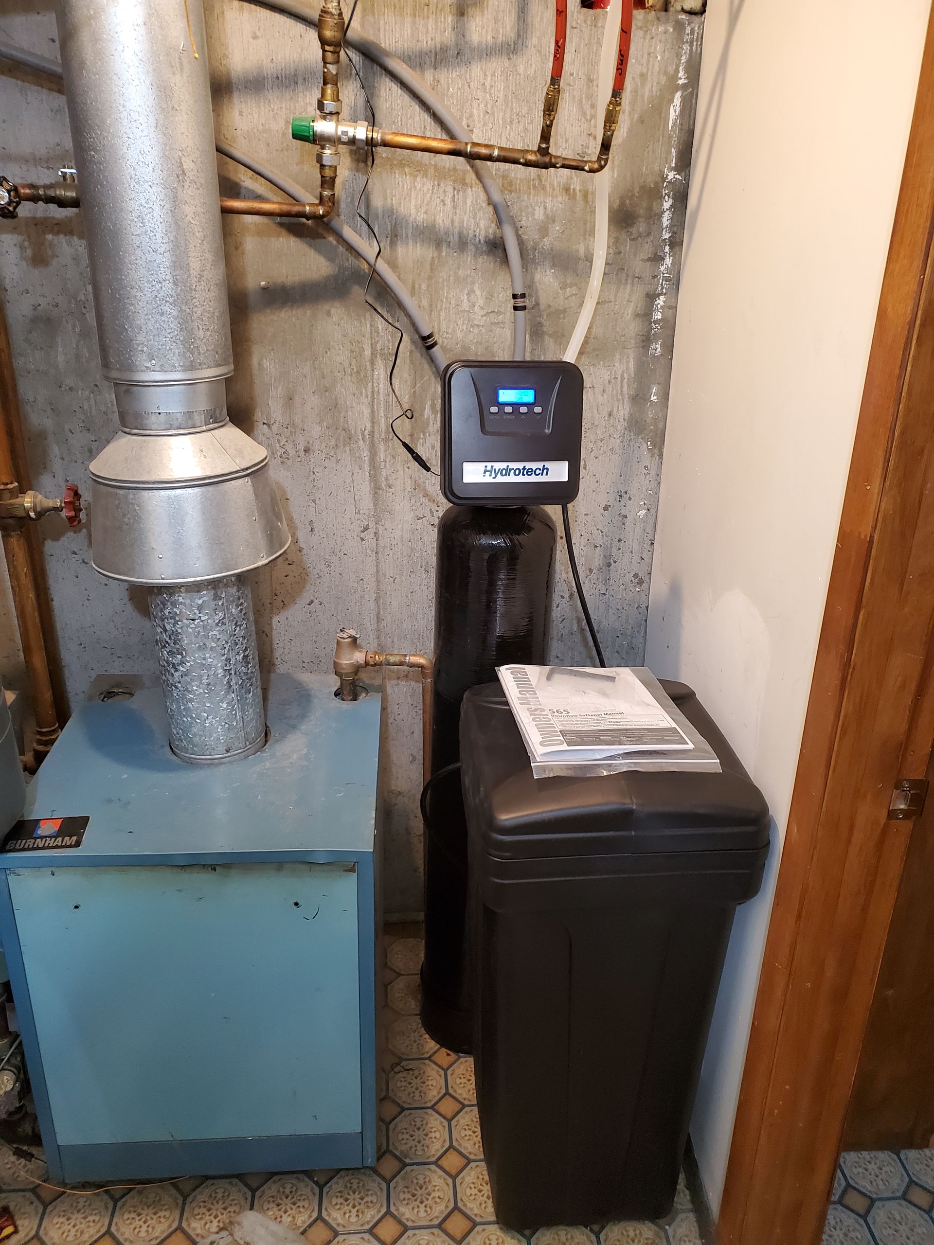 Water softener, water heater, and piping in a basement. The softener has a black tank and control panel.