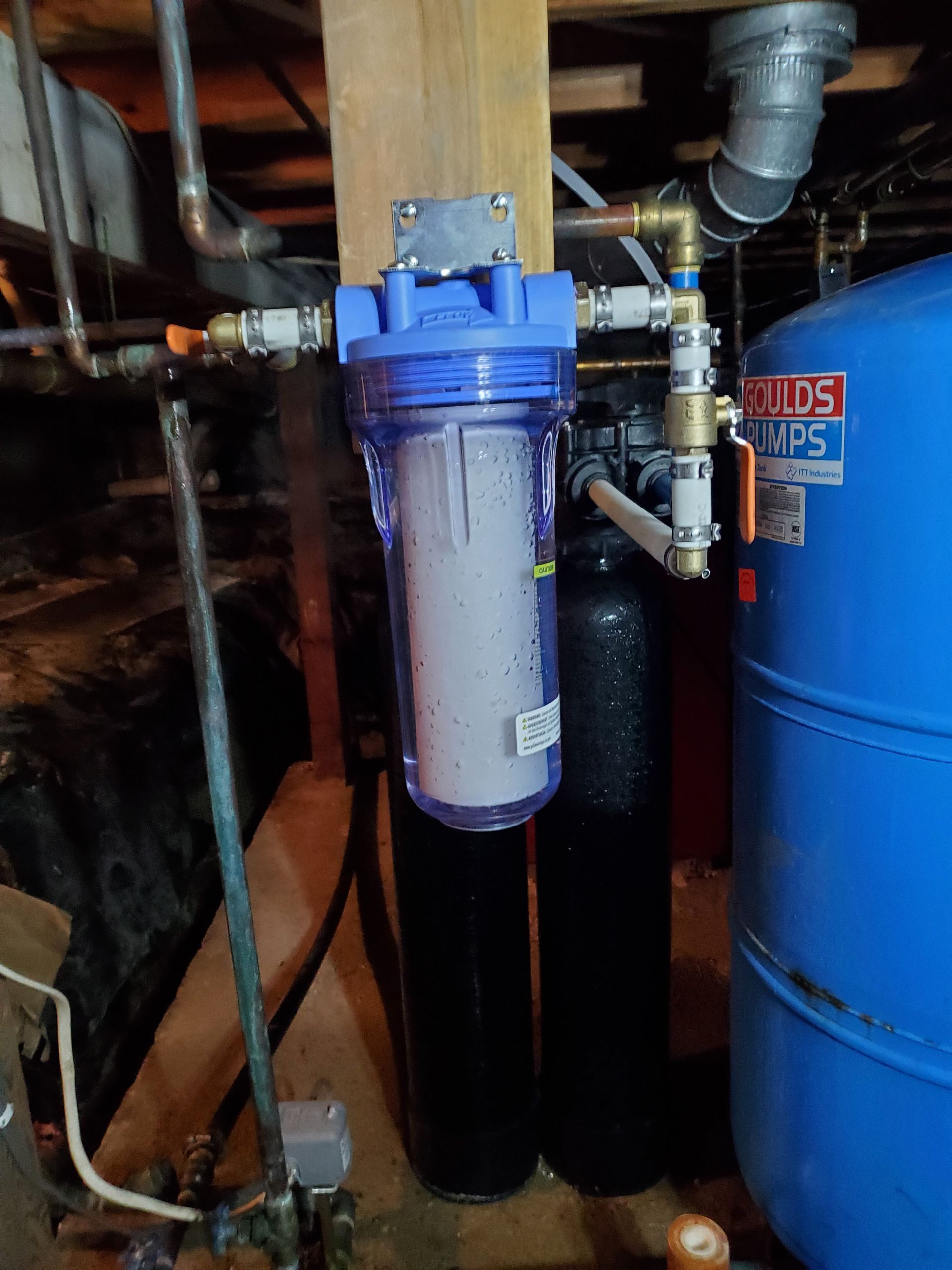 Water filtration system: clear filter housing, black tanks, pipes, and a blue water tank in a basement setting.